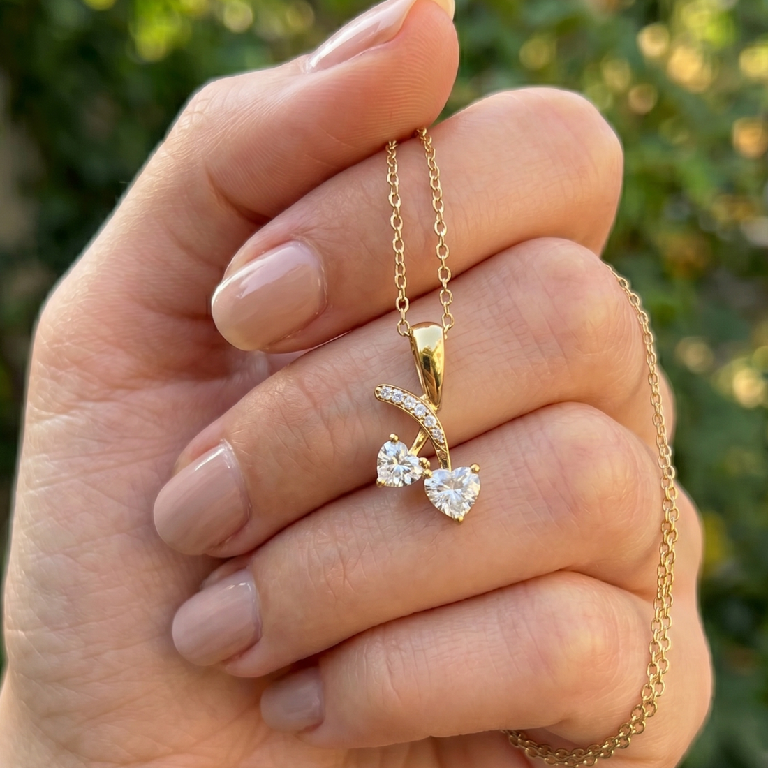 A close-up of a hand holding a delicate gold necklace with a leaf-shaped pendant set with two clear heart-shaped gemstones, against a blurred green outdoor background.