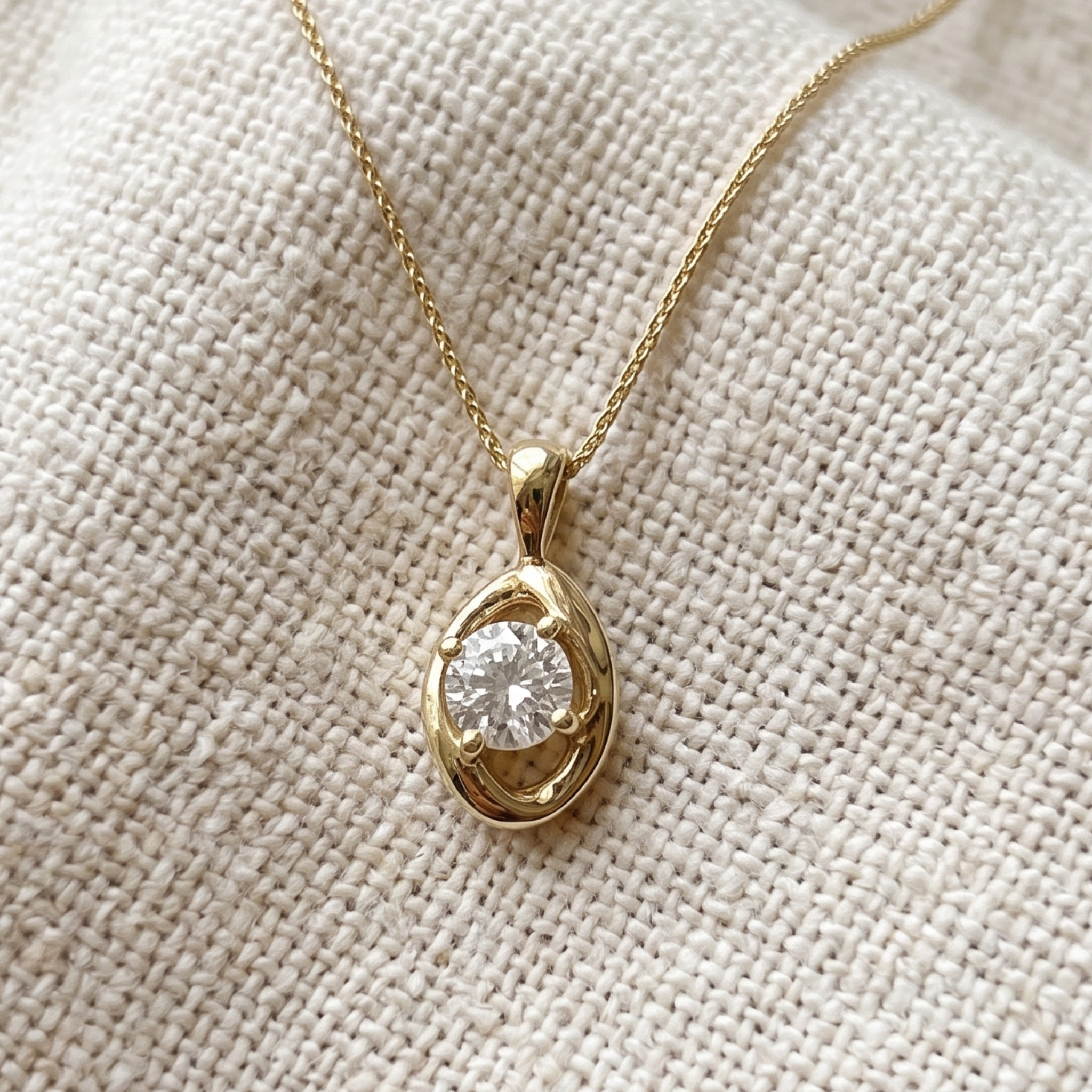 A gold pendant necklace with a round clear gemstone set in an organic, swirling gold bezel, displayed on a textured beige fabric background.