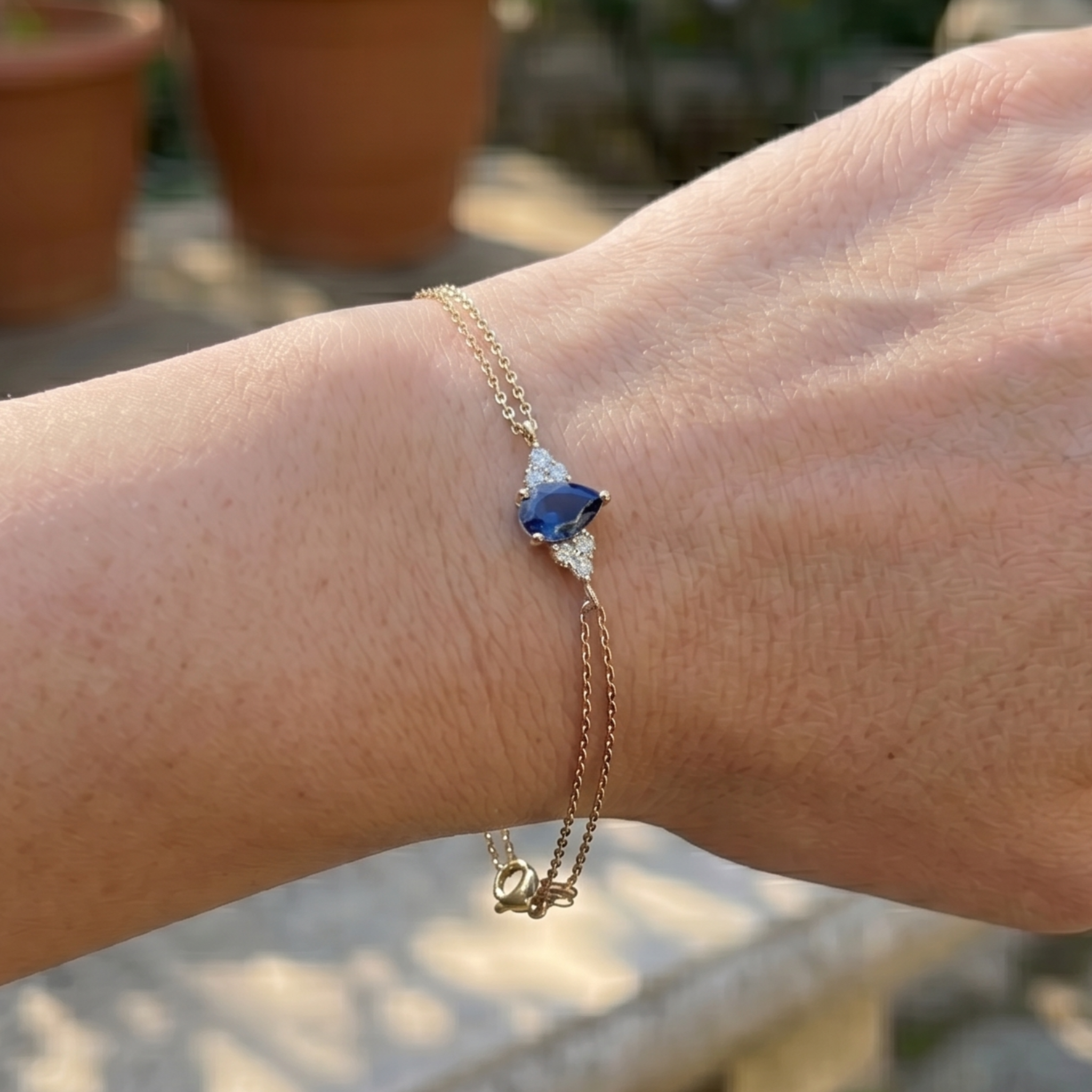 A delicate gold chain bracelet with a central blue and white triangular gemstone charm, shown worn on a person's wrist against a blurred outdoor background.