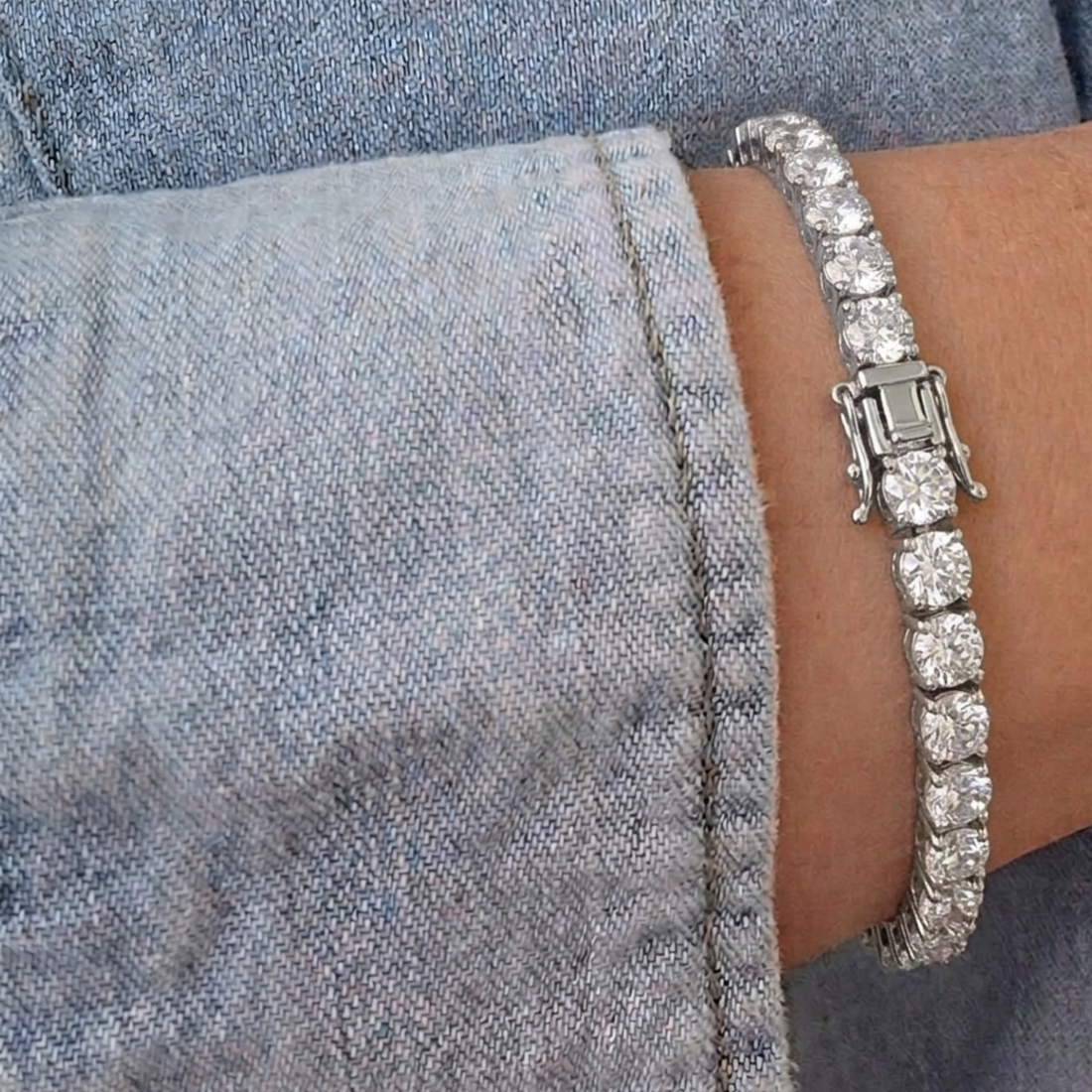 A silver-toned tennis bracelet with clear rectangular gemstones worn on a wrist, shown against a denim shirt background.