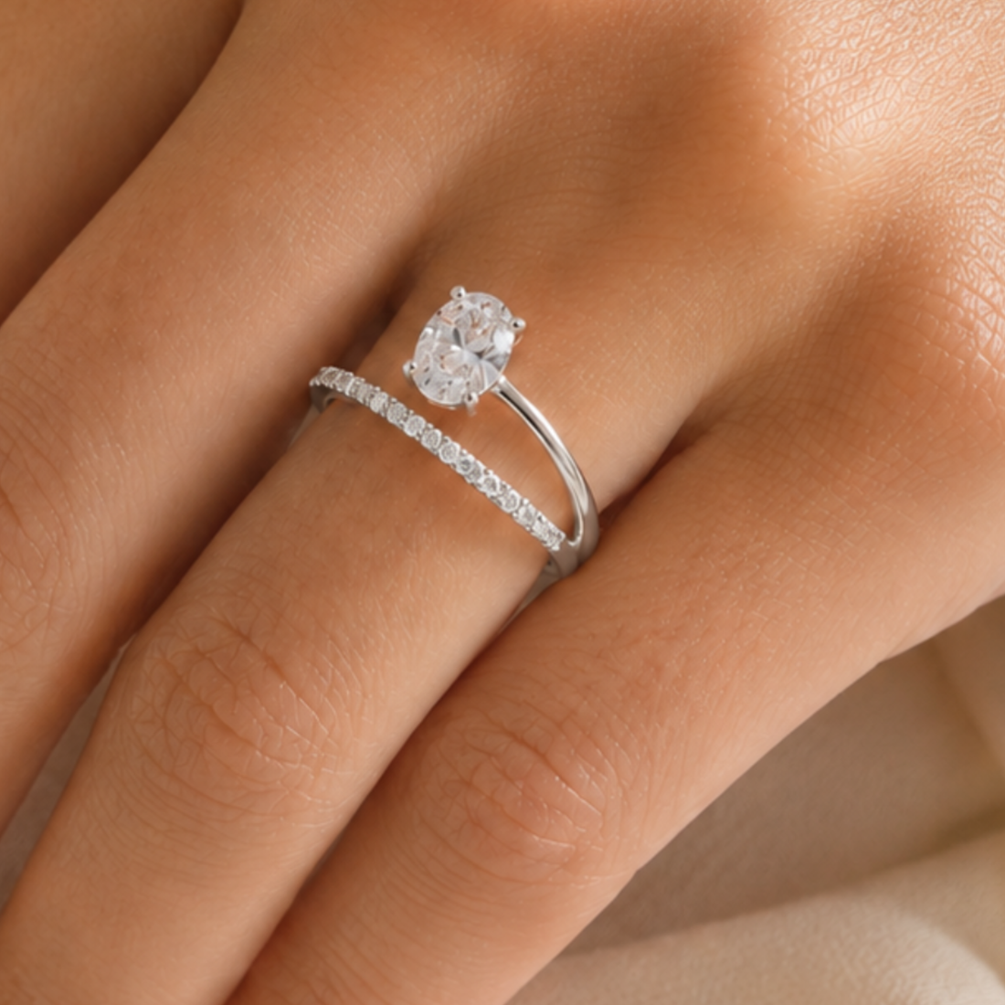 A close-up of a silver engagement ring with a round-cut clear center stone and a thin pavé band, worn on a finger.