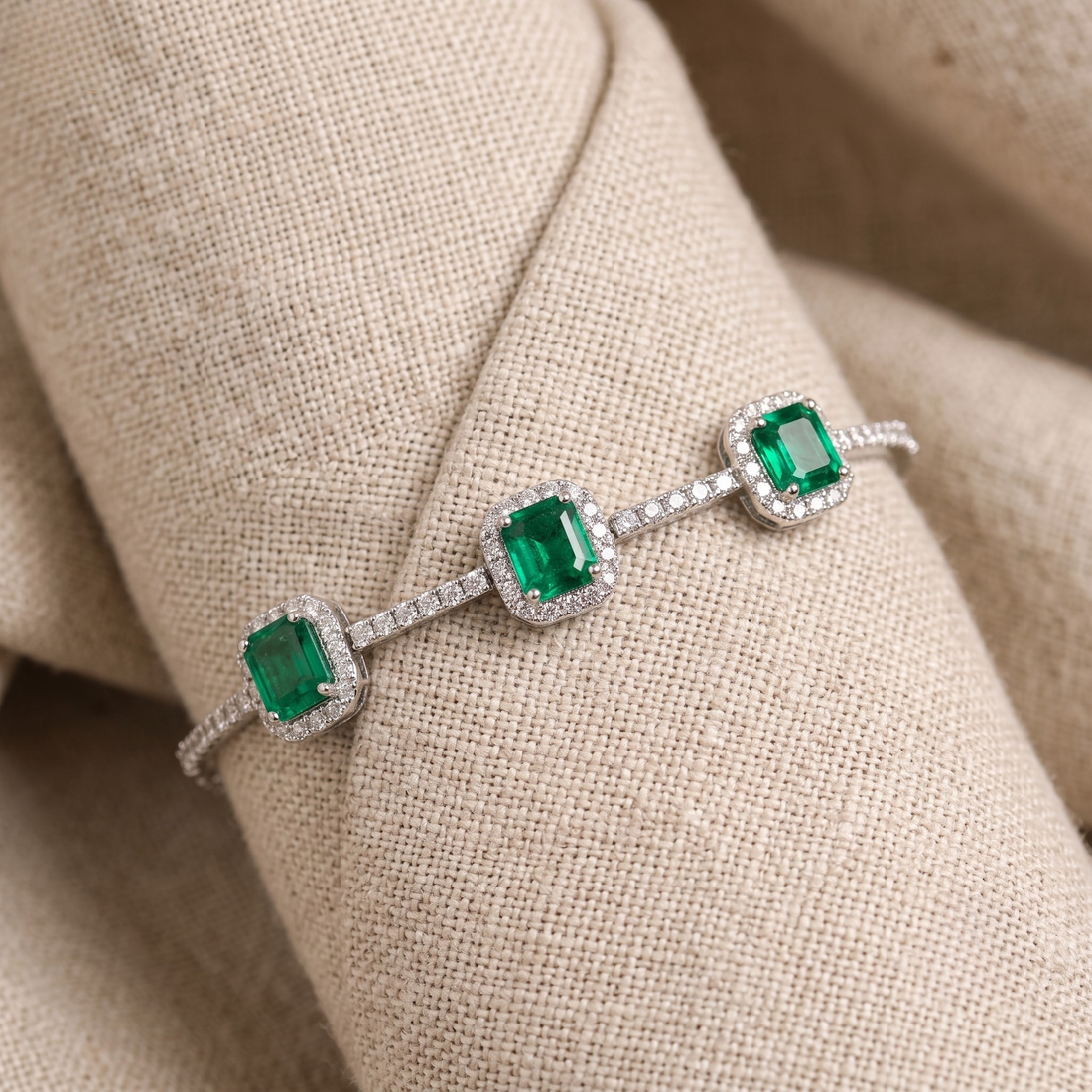 A silver-toned bracelet with three emerald-cut green gemstones, each surrounded by a halo of small clear stones, displayed on a beige fabric ring stand.