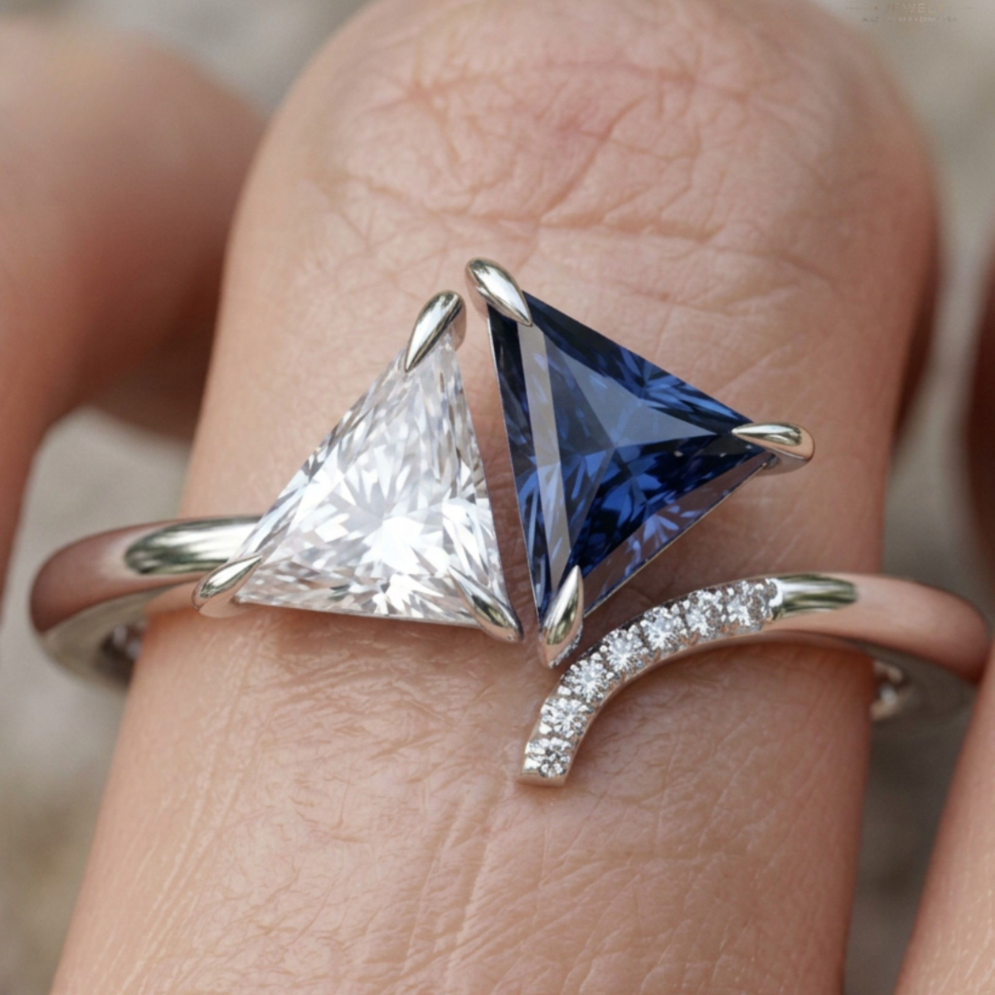 A close-up of a ring worn on a finger, featuring two triangular gemstones in silver settings—one clear and one deep blue—along with a curved row of small clear stones on a rose-gold-toned band.