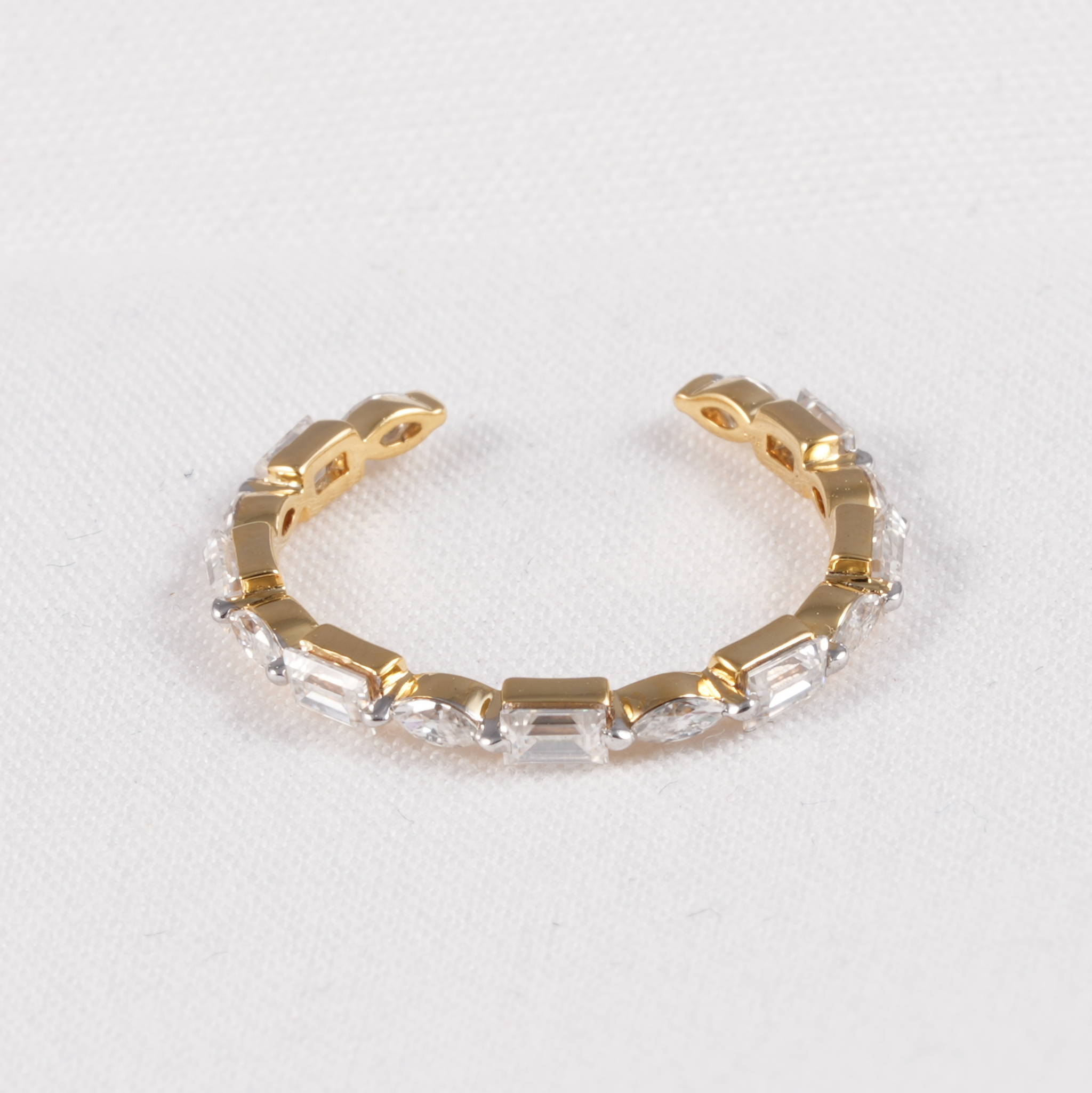 A gold-toned open cuff bracelet featuring a row of clear baguette and marquise-cut crystals, displayed on a textured white background.