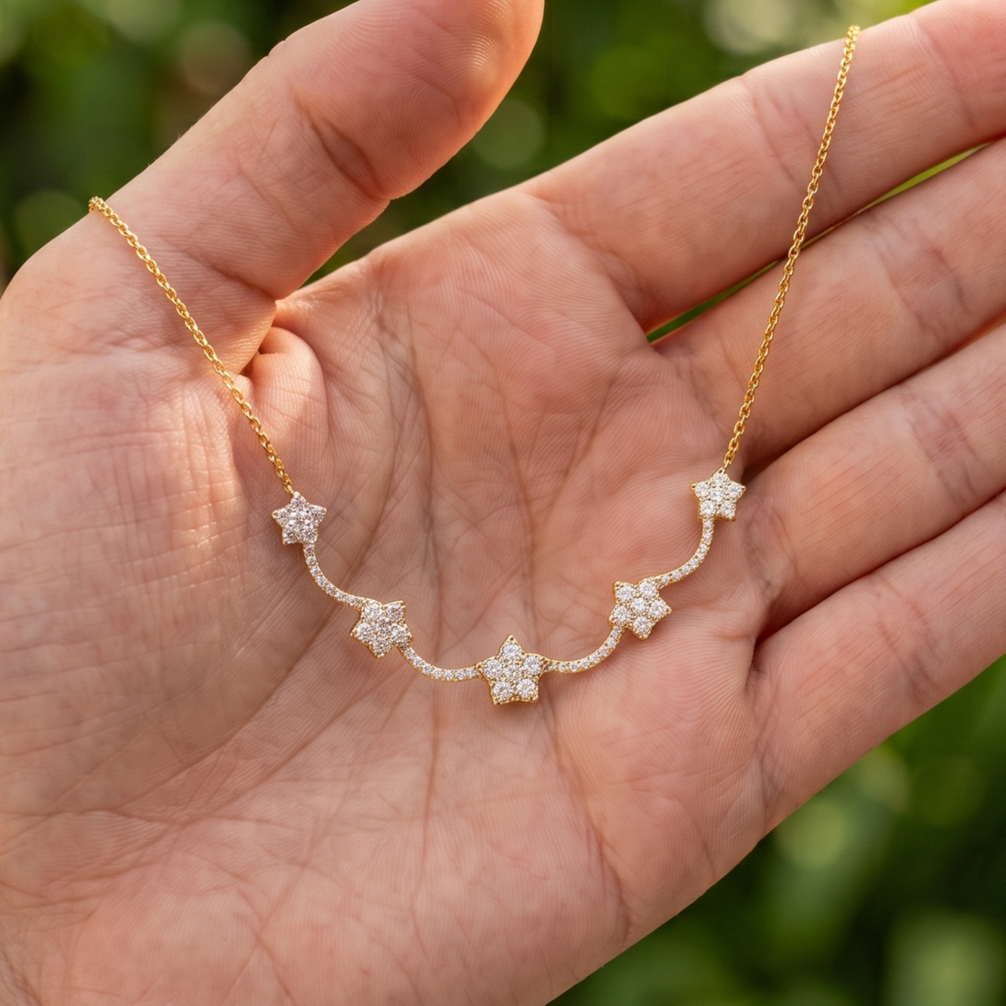 A delicate gold chain necklace with five star-shaped puffed pendant clusters set with small clear stones, held in the palm of a hand against a blurred green background.