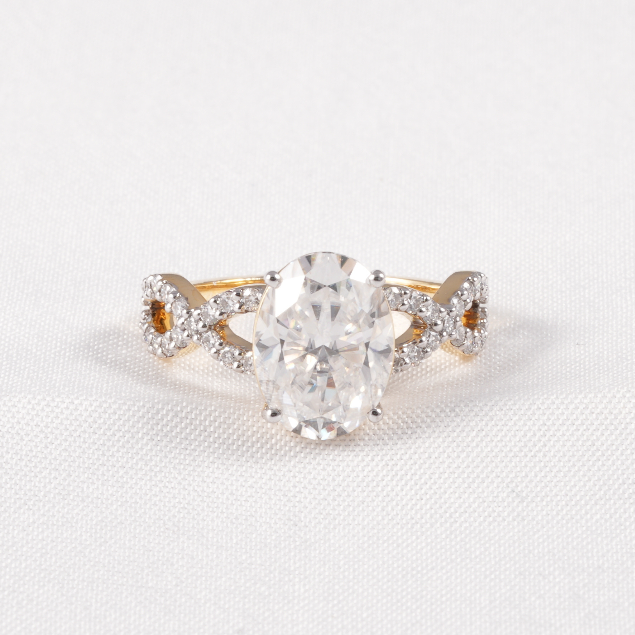A yellow gold engagement ring featuring a large oval-cut clear center stone and pavé-set accent stones on the band, photographed on a textured white fabric background.