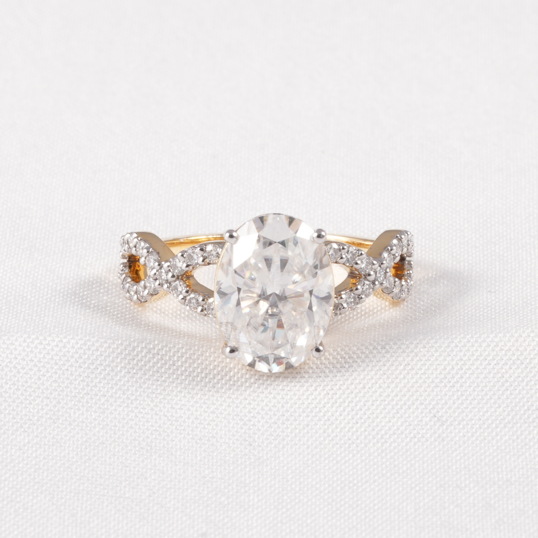 A yellow gold engagement ring featuring a large oval-cut clear center stone and pavé-set accent stones on the band, photographed on a textured white fabric background.