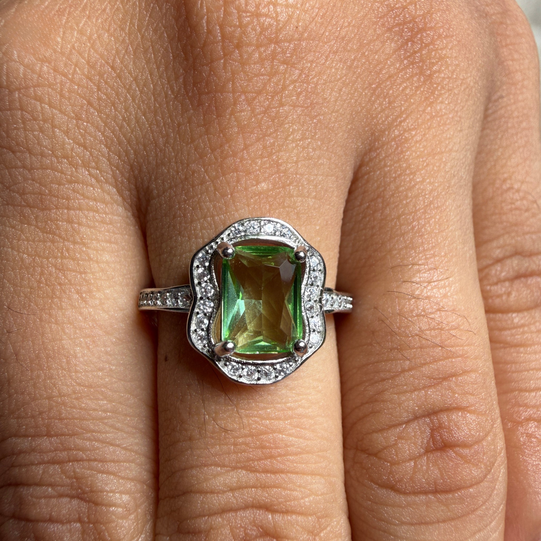 A close-up of a ring worn on a finger, featuring a large rectangular green gemstone with a scalloped silver halo and small accent stones on the band.