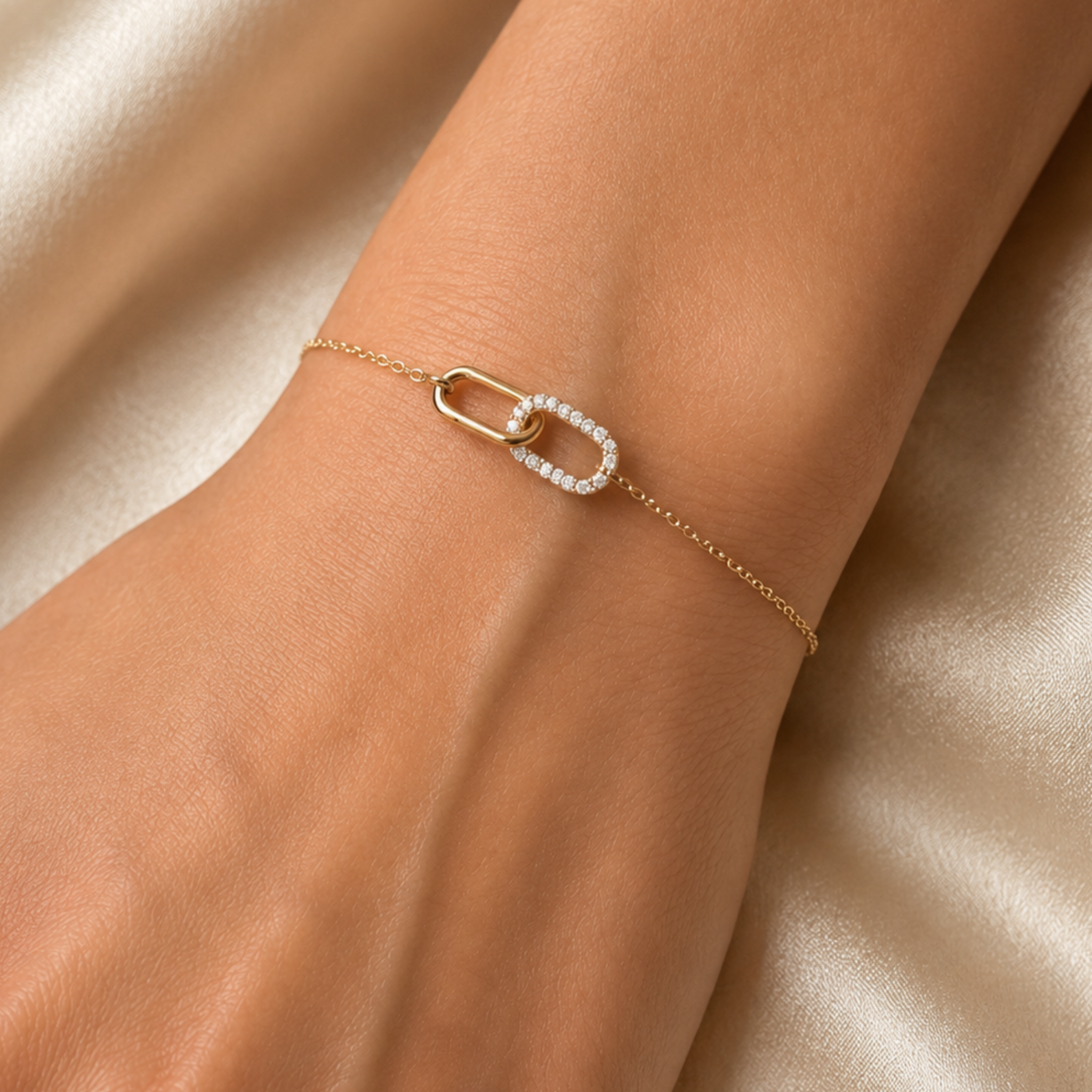 A delicate gold chain bracelet with a central open-link design, one side of which is lined with small clear stones, shown worn on a wrist against a soft beige satin background.