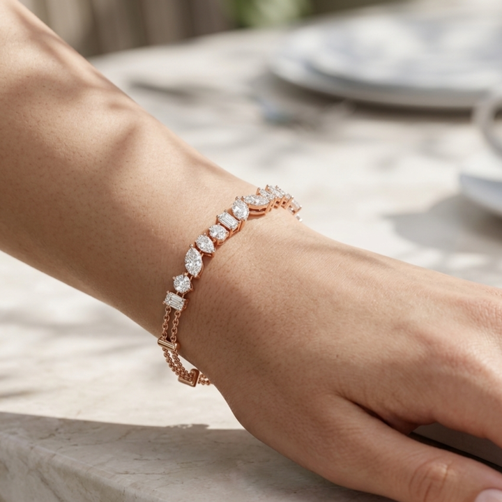 A close-up of a person's wrist wearing a rose gold bracelet with marquise-cut clear gemstones, set against a softly blurred outdoor table background.