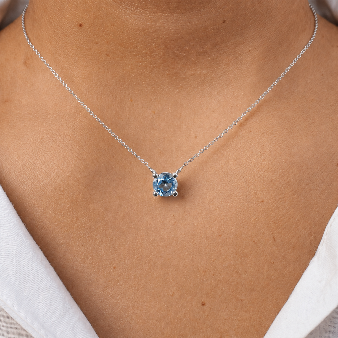A close-up of a person's neck and chest wearing a delicate silver chain necklace with a single round-cut light blue gemstone pendant.