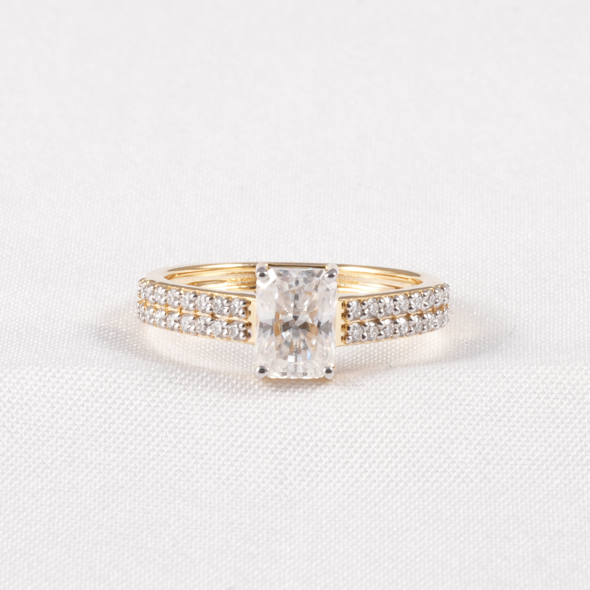 A yellow gold engagement ring featuring a rectangular radiant-cut diamond center stone and pavé-set diamonds along the band, displayed on a textured white fabric background.