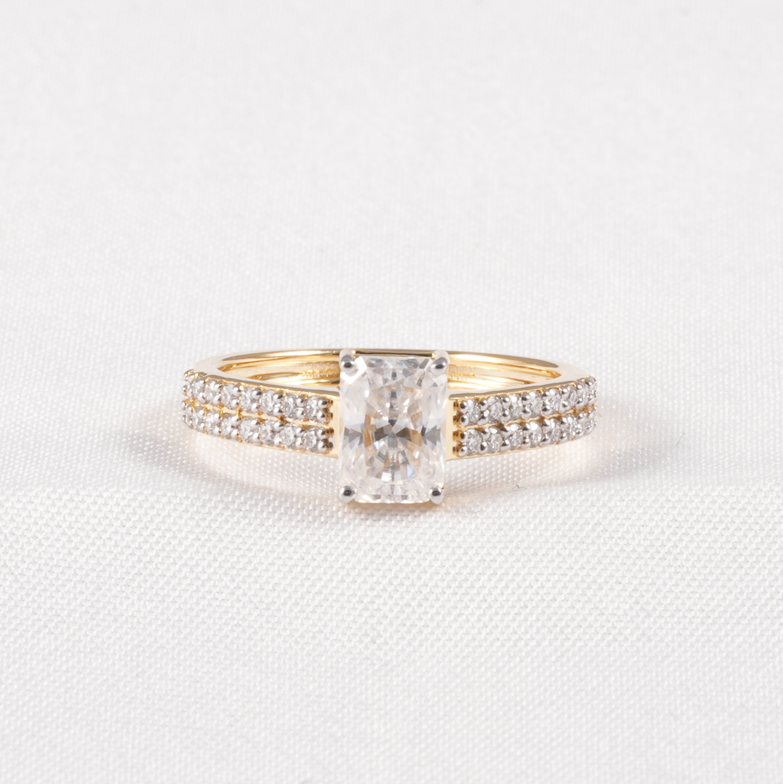A yellow gold engagement ring featuring a rectangular radiant-cut diamond center stone and pavé-set diamonds along the band, displayed on a textured white fabric background.