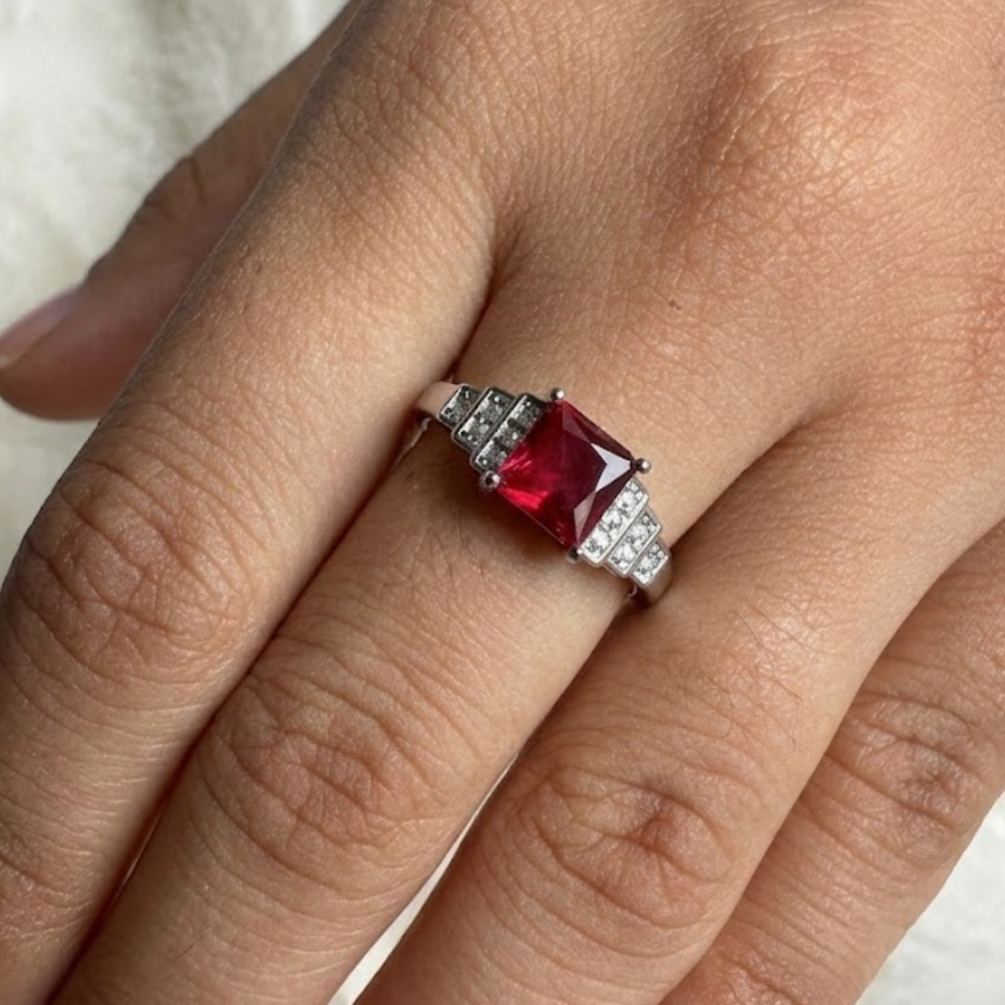 A close-up of a hand wearing a silver ring with a square-cut red gemstone and small clear accent stones on the band.