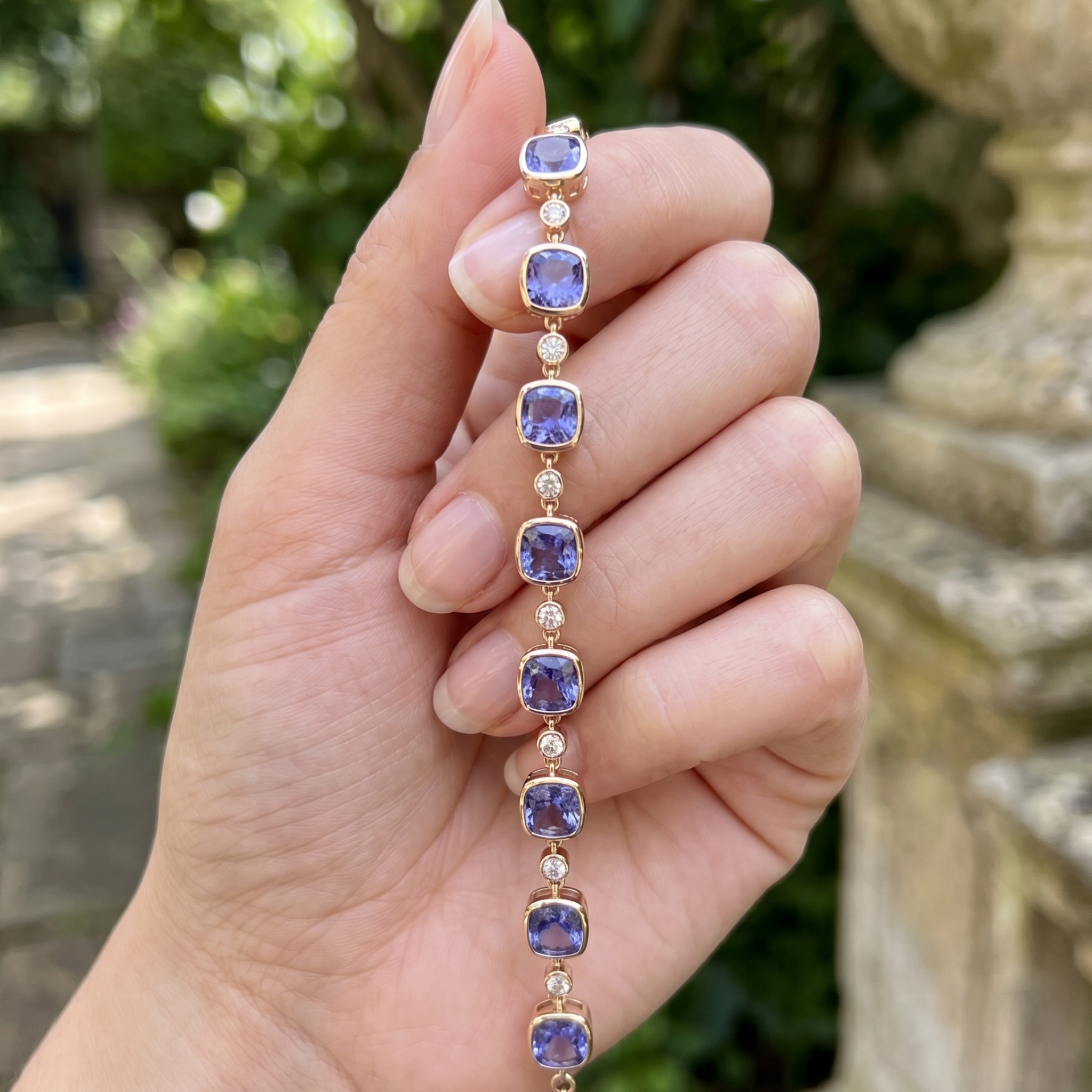 A hand holding a gold bracelet with square purple tanzanite stones and small clear accent stones, photographed outdoors with a blurred garden background.