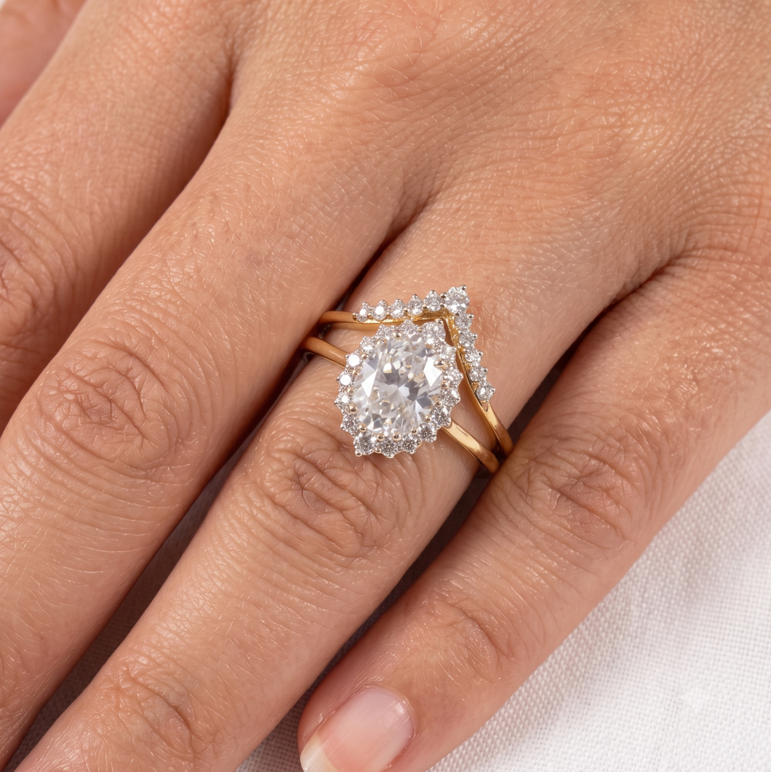 A close-up of a hand wearing a gold ring set with a large pear-cut clear gemstone and smaller surrounding stones, with a matching slim gold band visible behind it.