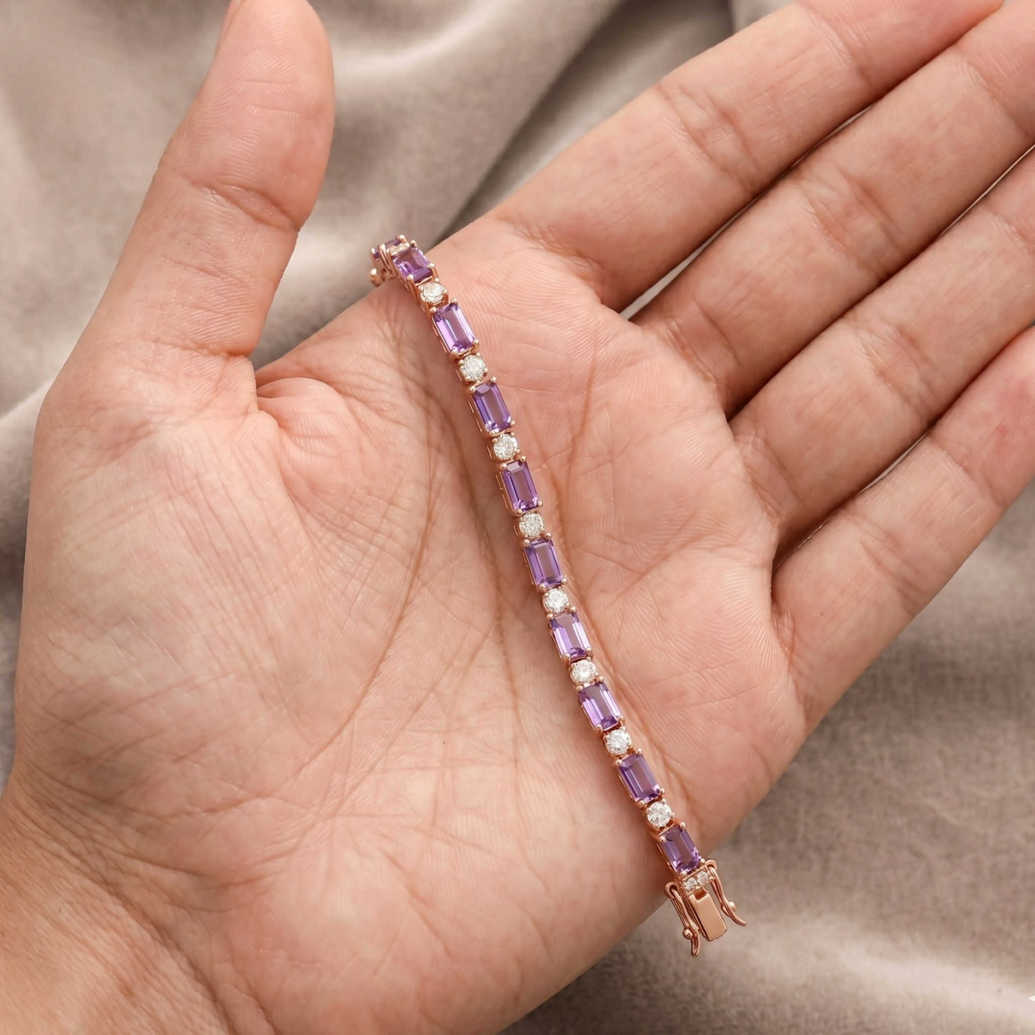 A rose gold tennis bracelet with purple and white stones, held in a person's hand against a soft beige fabric background.