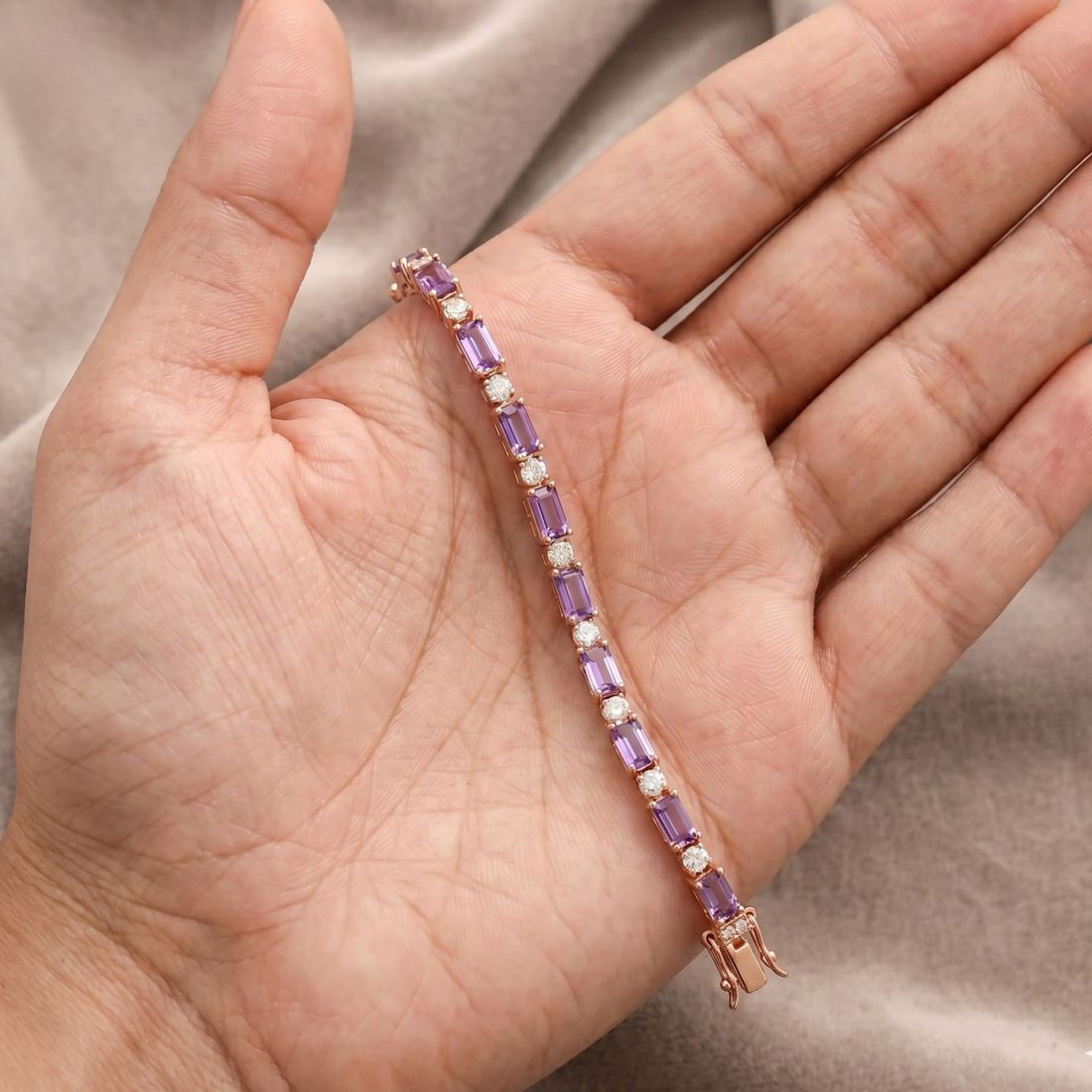A rose gold tennis bracelet with purple and white stones, held in a person's hand against a soft beige fabric background.