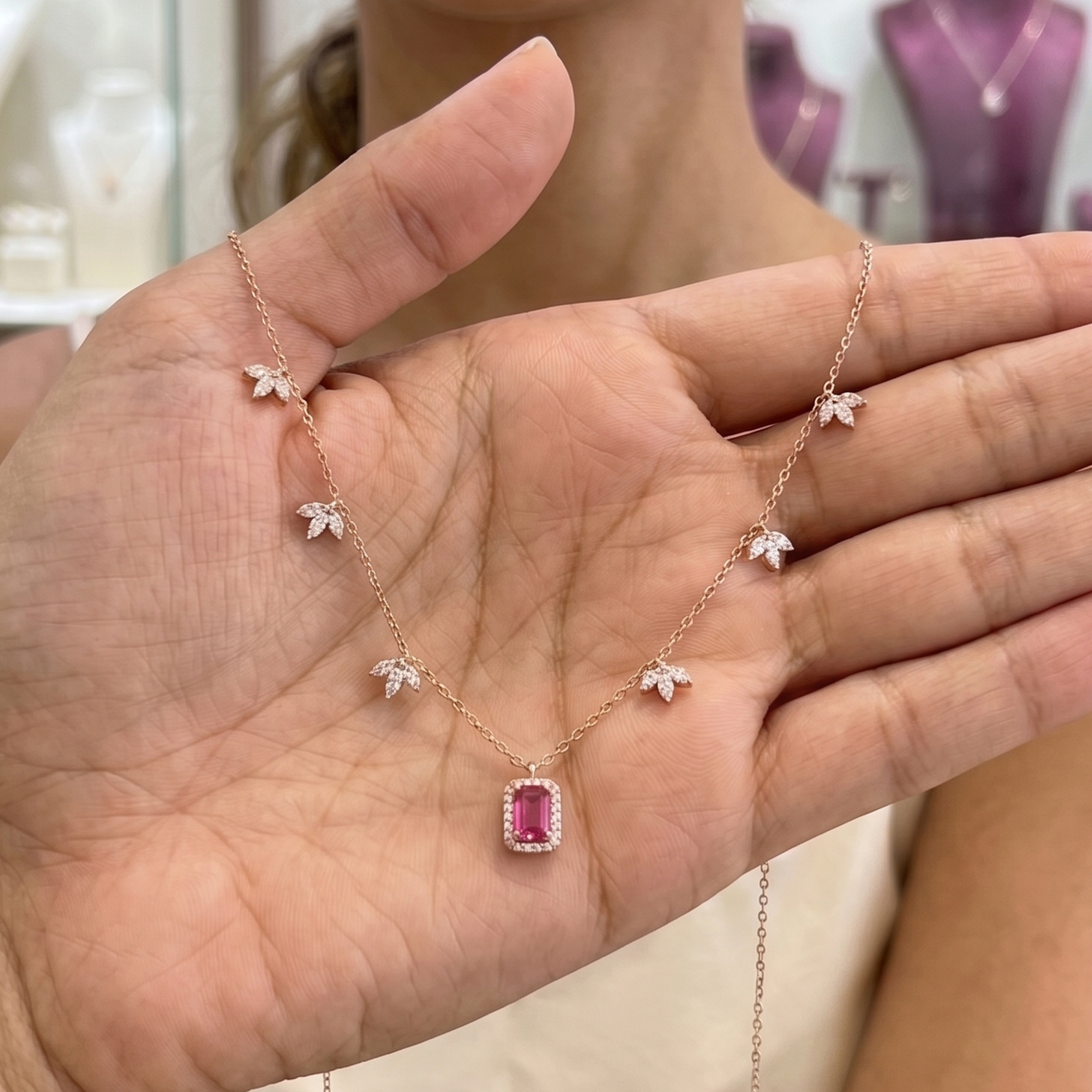 A rose gold necklace with a rectangular pink gemstone pendant and small flower motifs, held in a person's hand.