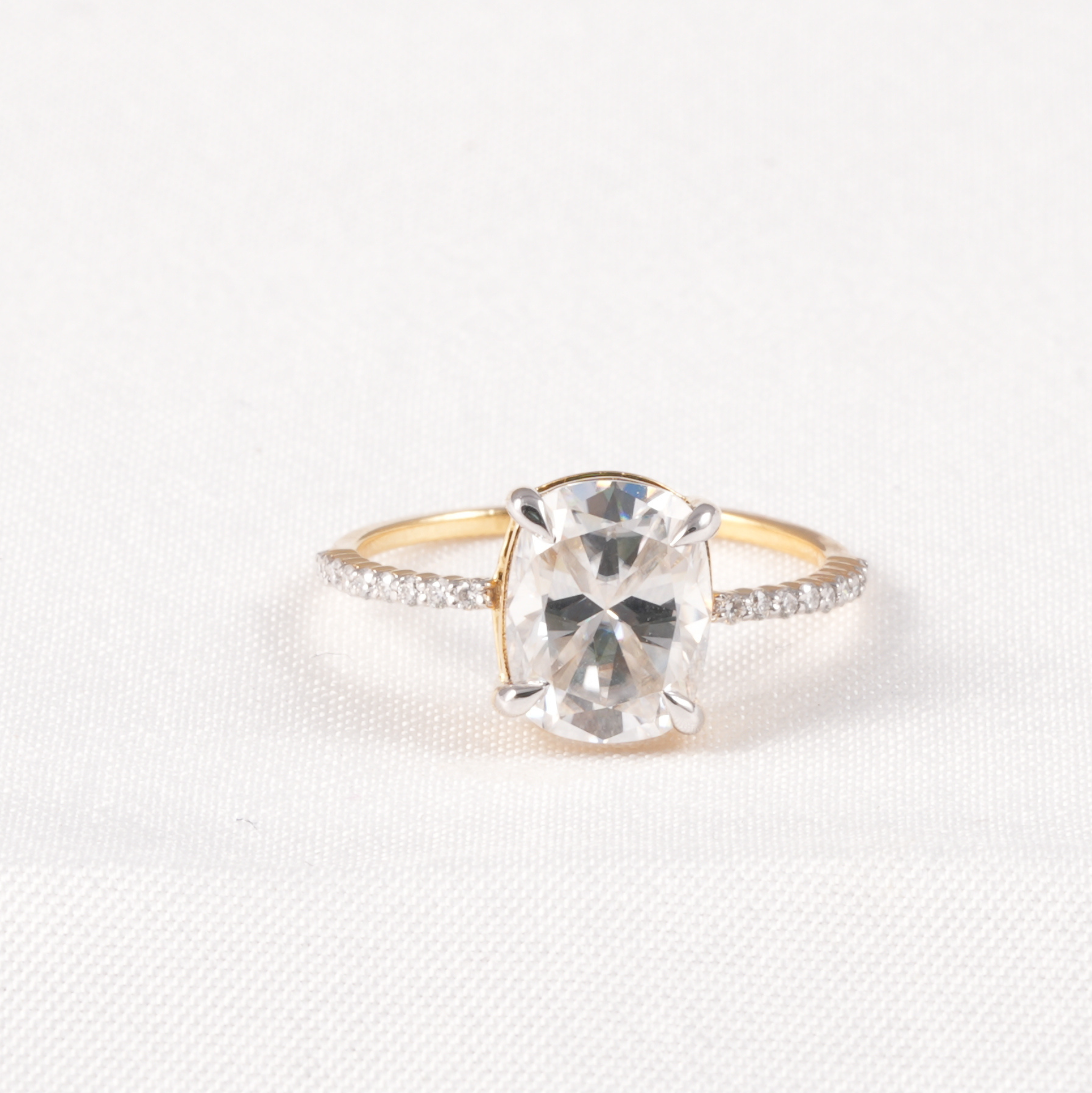 A yellow gold ring with a large oval clear gemstone in a prong setting, accented by small pavé diamonds along the shoulders, shown on a textured white background.