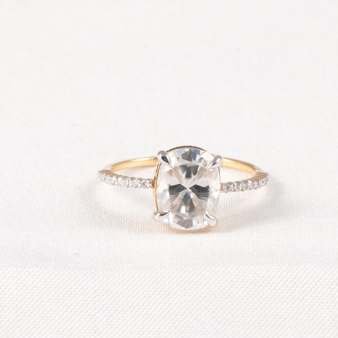 A yellow gold ring with a large oval clear gemstone in a prong setting, accented by small pavé diamonds along the shoulders, shown on a textured white background.