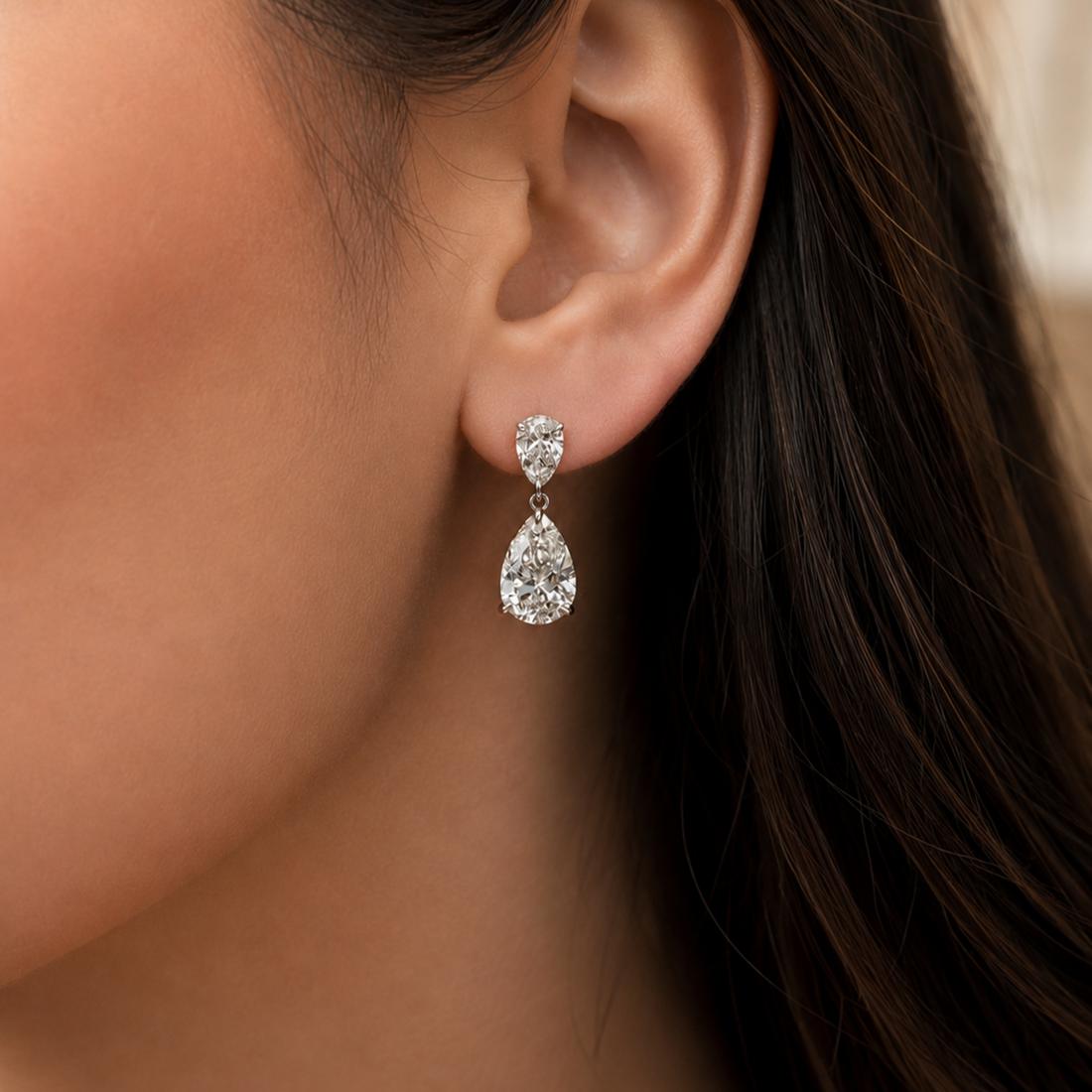A close-up of a woman's ear wearing a silver-tone teardrop dangle earring with a clear teardrop-cut gemstone and a smaller round-cut stone above it.