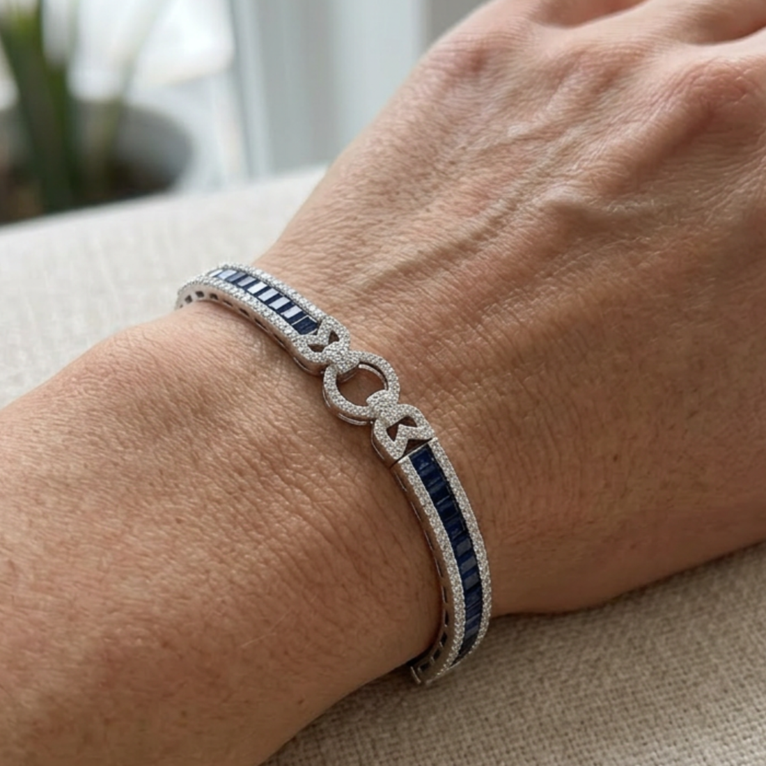 A close-up of a person's wrist wearing a silver-toned bracelet with a double interlocking rings motif paved with small clear stones, flanked by vertical dark blue inlay segments.