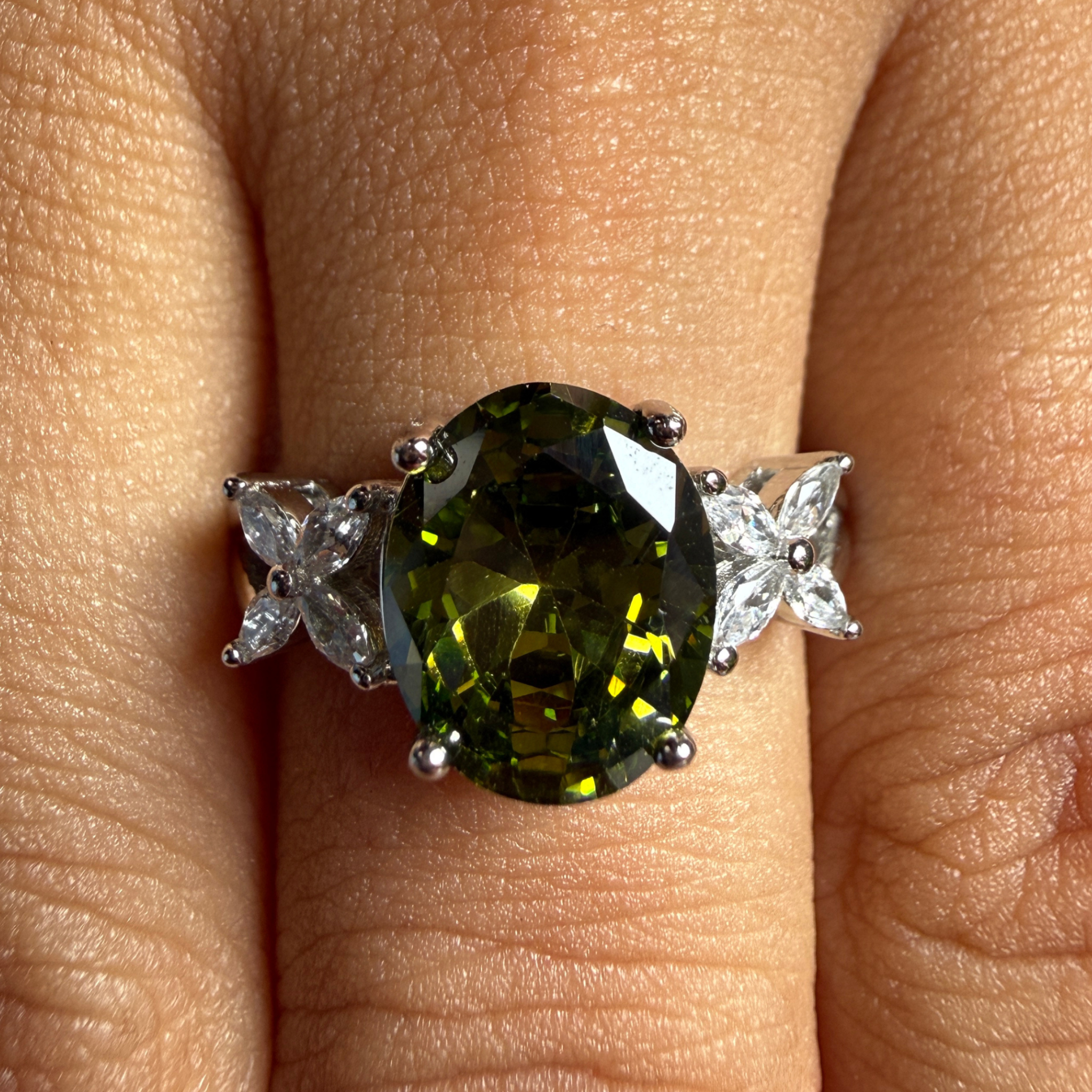 An oval-cut olive green gemstone ring with silver-colored metal settings and clear accent stones, shown worn on a finger.