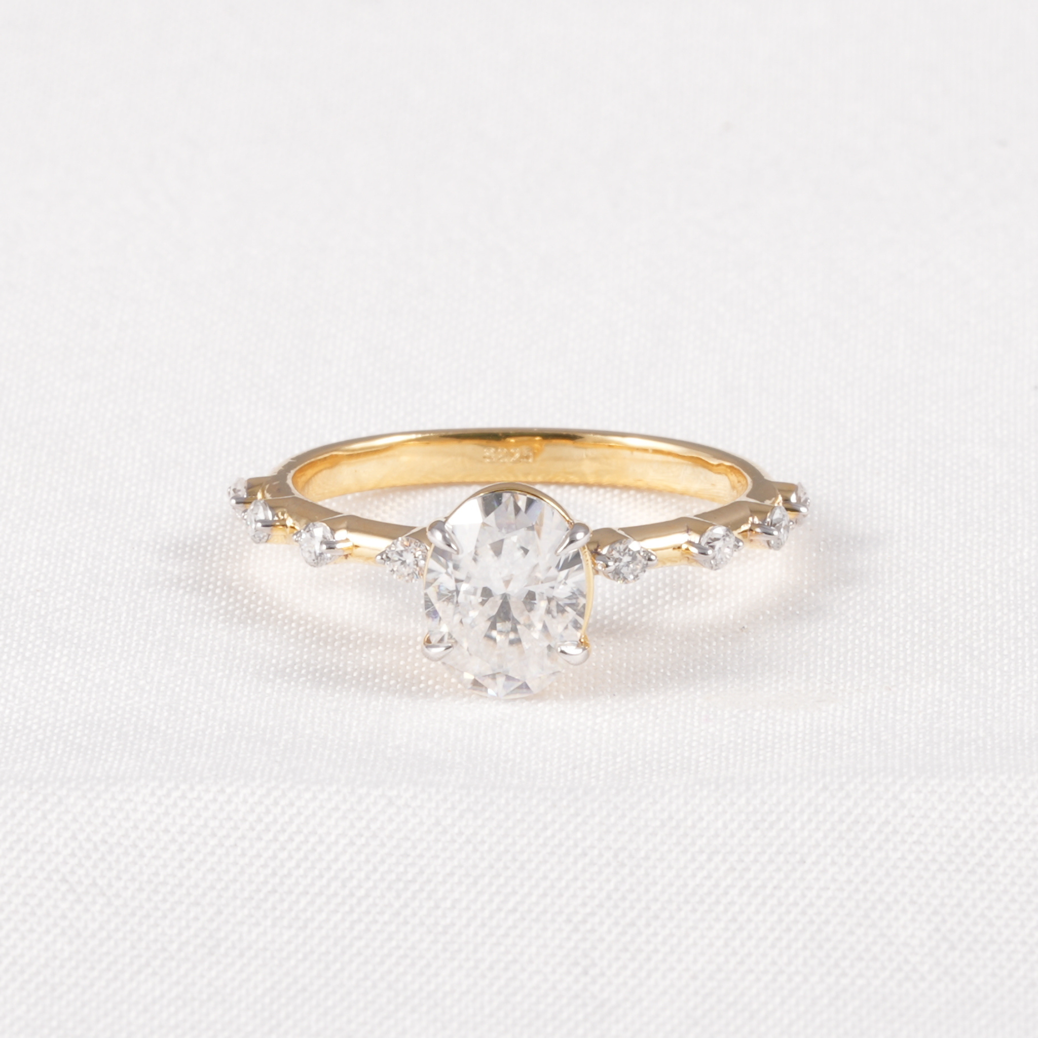 A yellow gold ring featuring a large oval-cut clear center stone and smaller accent stones along the band, set against a white textured fabric background.