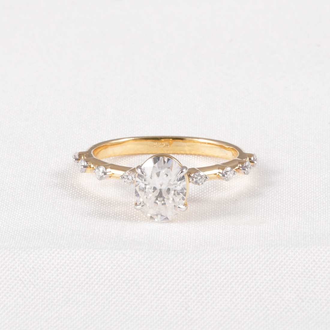 A yellow gold ring featuring a large oval-cut clear center stone and smaller accent stones along the band, set against a white textured fabric background.
