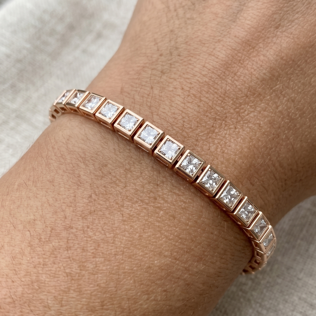 A rose gold tennis bracelet with square-cut clear gemstones, shown worn on a person's wrist against a neutral light beige fabric background.
