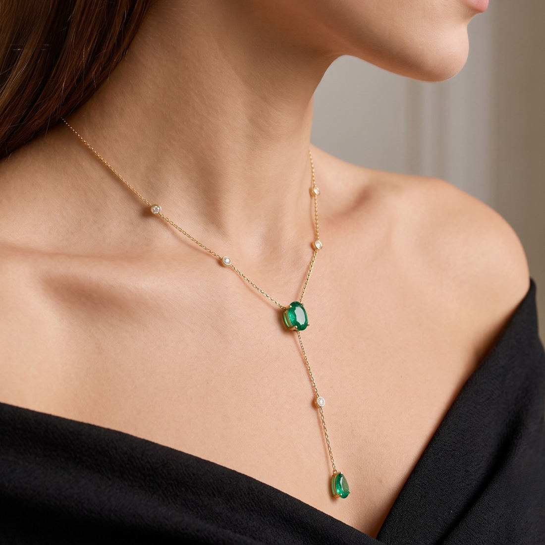 A close-up of a woman wearing a delicate gold Y-necklace with two green teardrop emerald-cut gemstones and small white bead accents.
