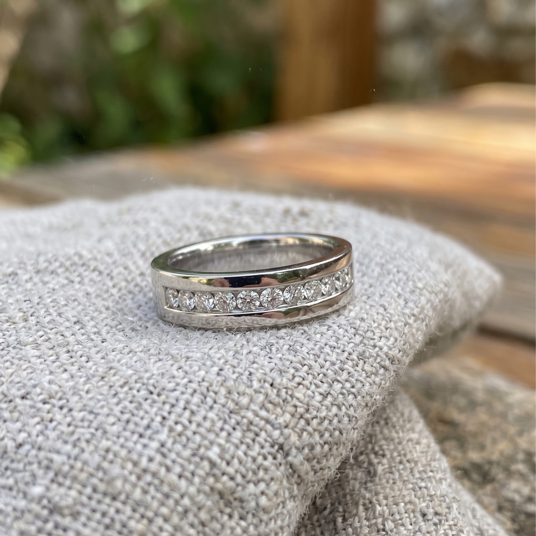 A silver band ring with a row of clear pavé gemstones, resting on a textured beige fabric surface with a blurred outdoor background.