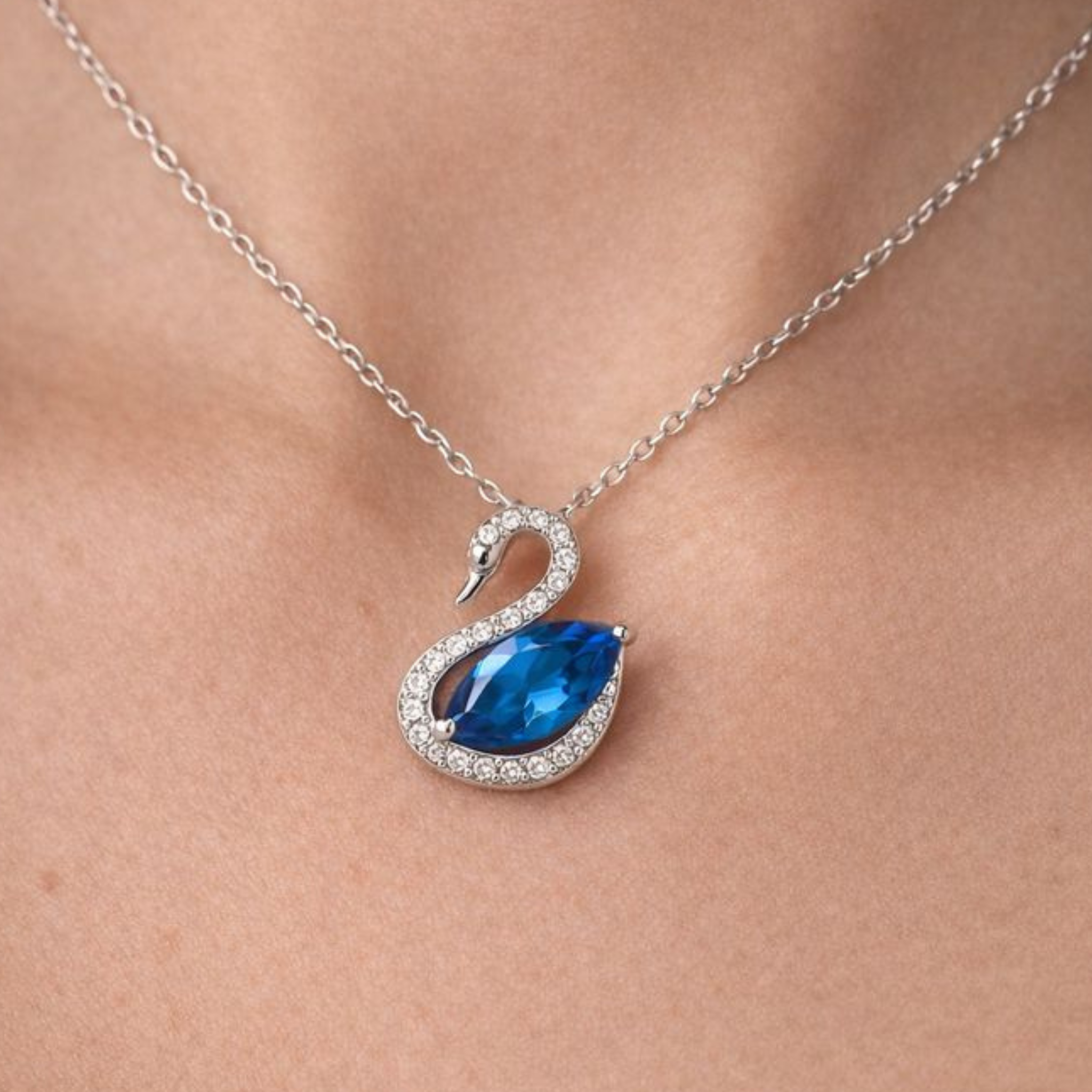 A silver-tone swan pendant necklace with a large blue teardrop gemstone and small clear stones, shown worn on a person's neck.