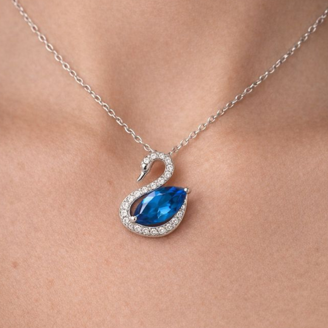 A silver-tone swan pendant necklace with a large blue teardrop gemstone and small clear stones, shown worn on a person's neck.