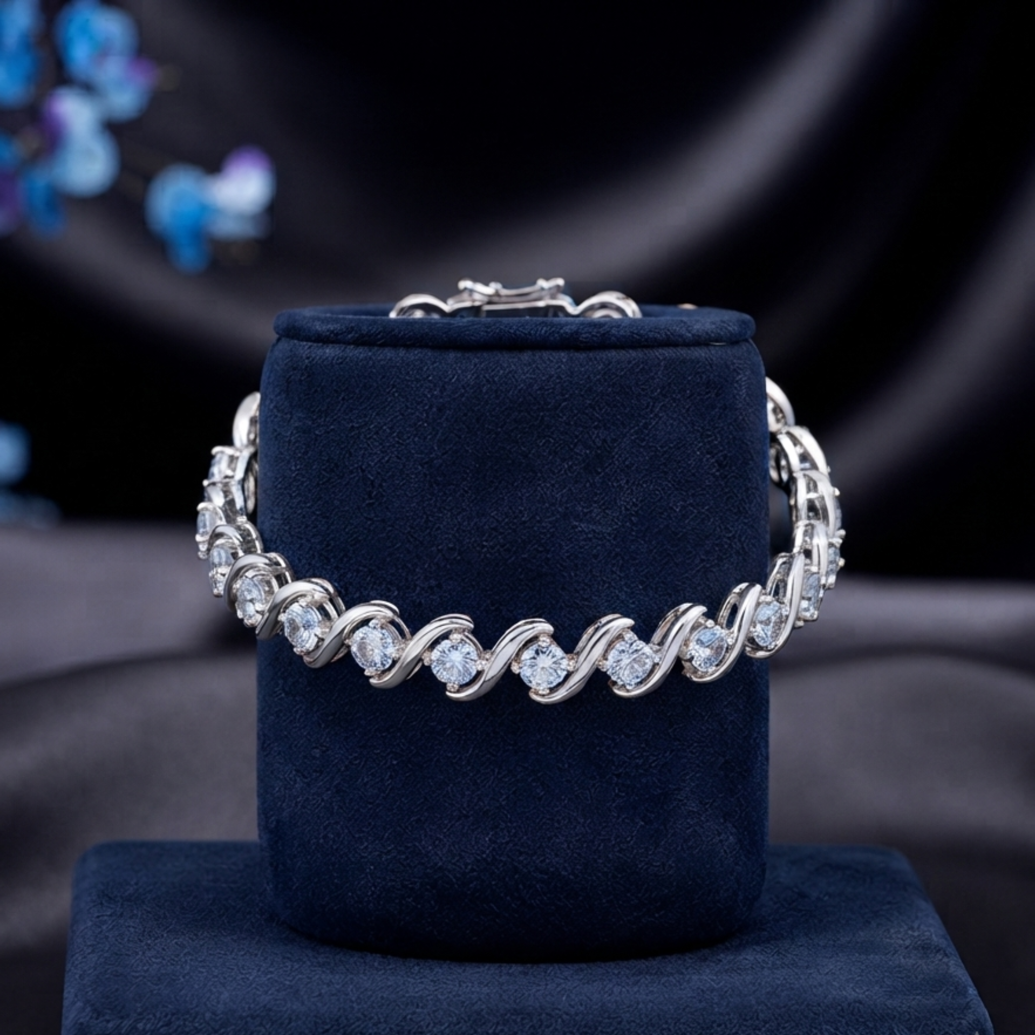A silver-toned tennis bracelet with clear round-cut stones, displayed on a dark navy velvet jewelry stand against a dark draped fabric background.