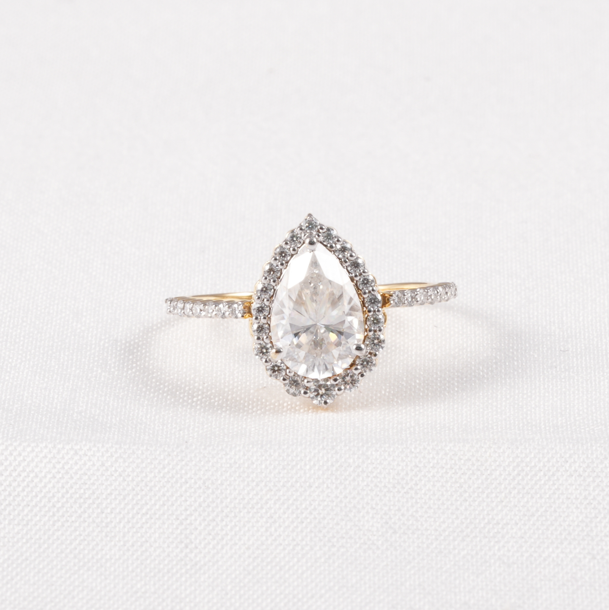 A pear-shaped halo ring featuring a clear central gemstone surrounded by a silver-toned halo and a yellow-gold band accented with small pavé stones, set against a clean white background.