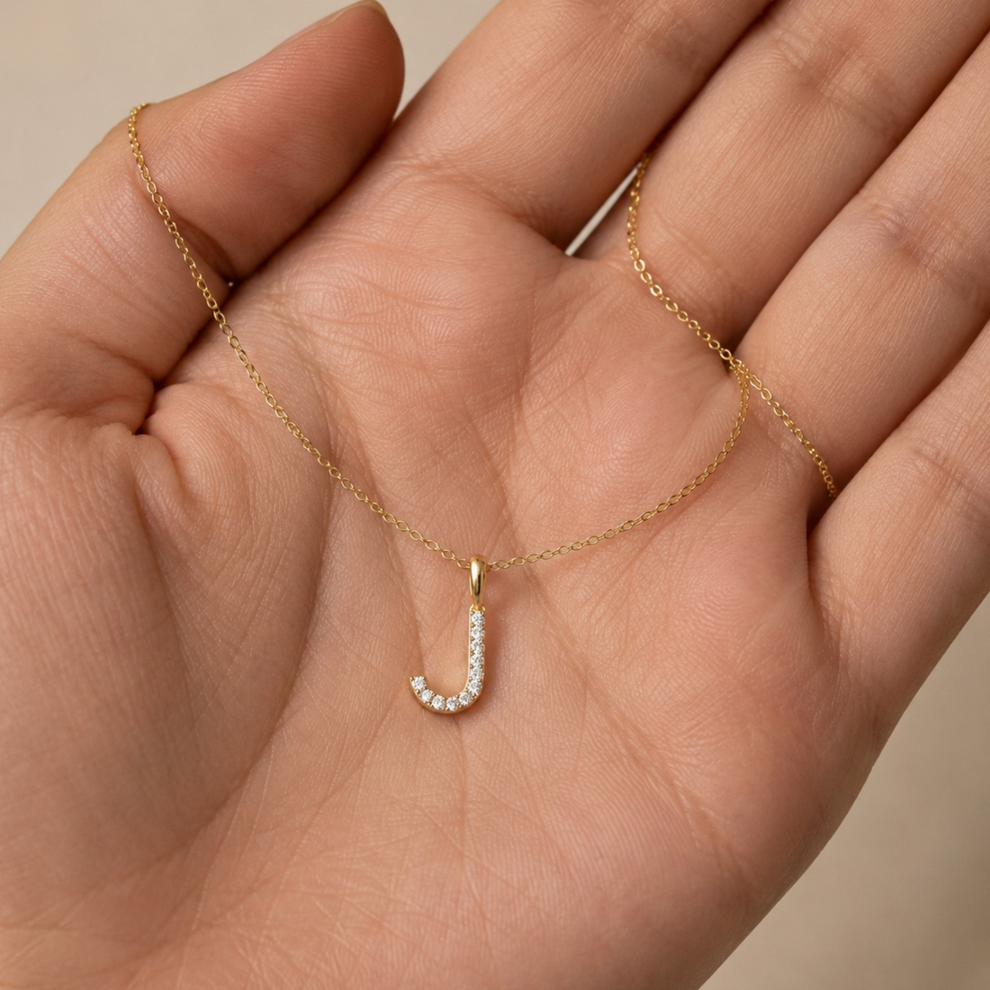 A delicate gold chain necklace with a small J-shaped pendant set with clear stones, held in the palm of a hand.