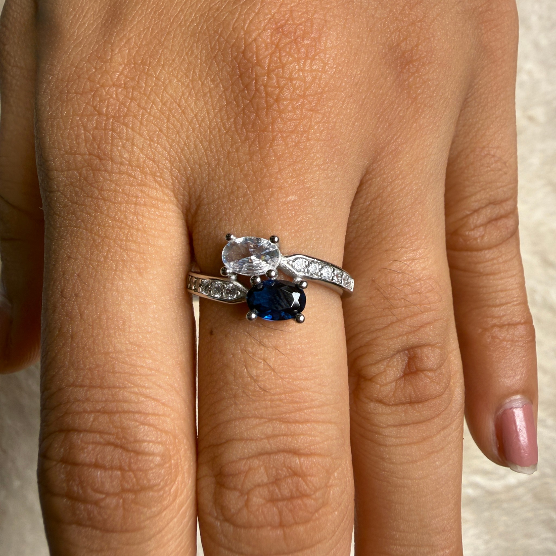 A close-up of a hand wearing a silver ring with a large clear oval gemstone and a smaller round blue sapphire stone, with small clear stones along the band.