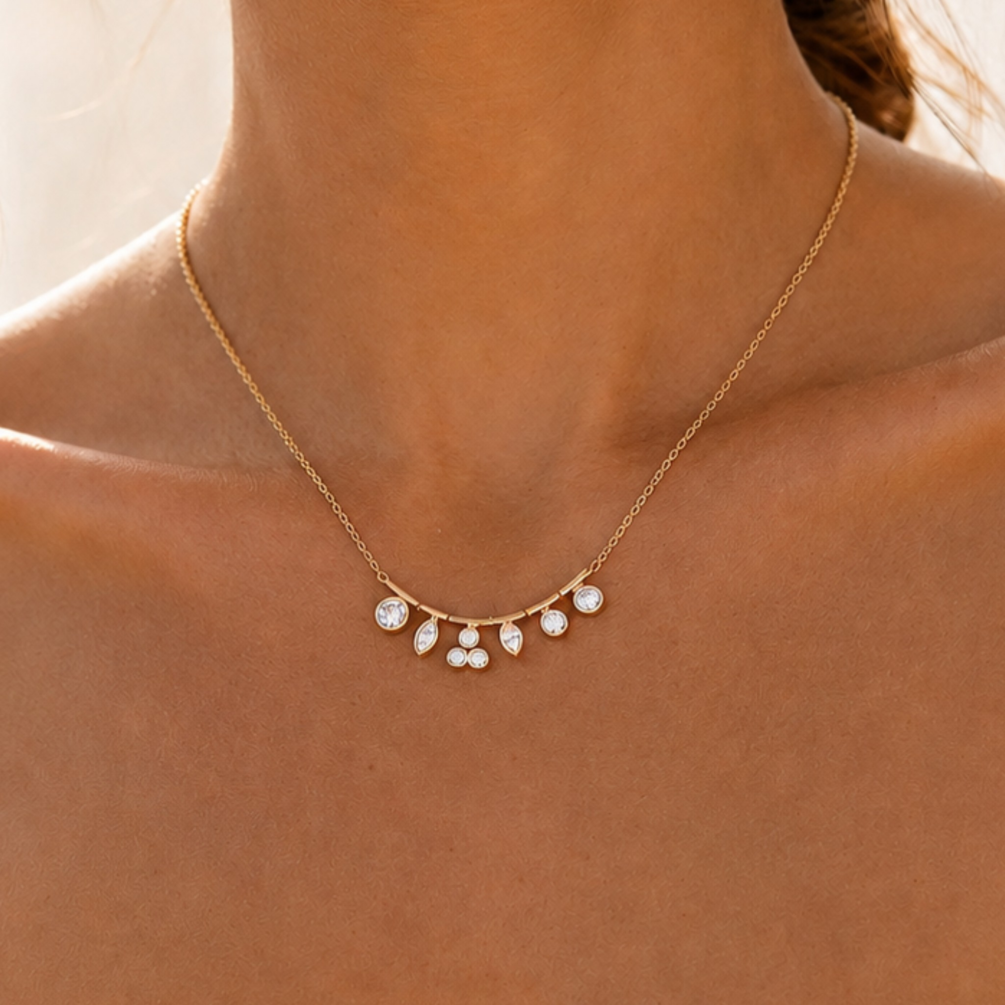 A delicate gold chain necklace with five small, clear teardrop-shaped stones arranged in a fan shape, shown worn on a model's neck.