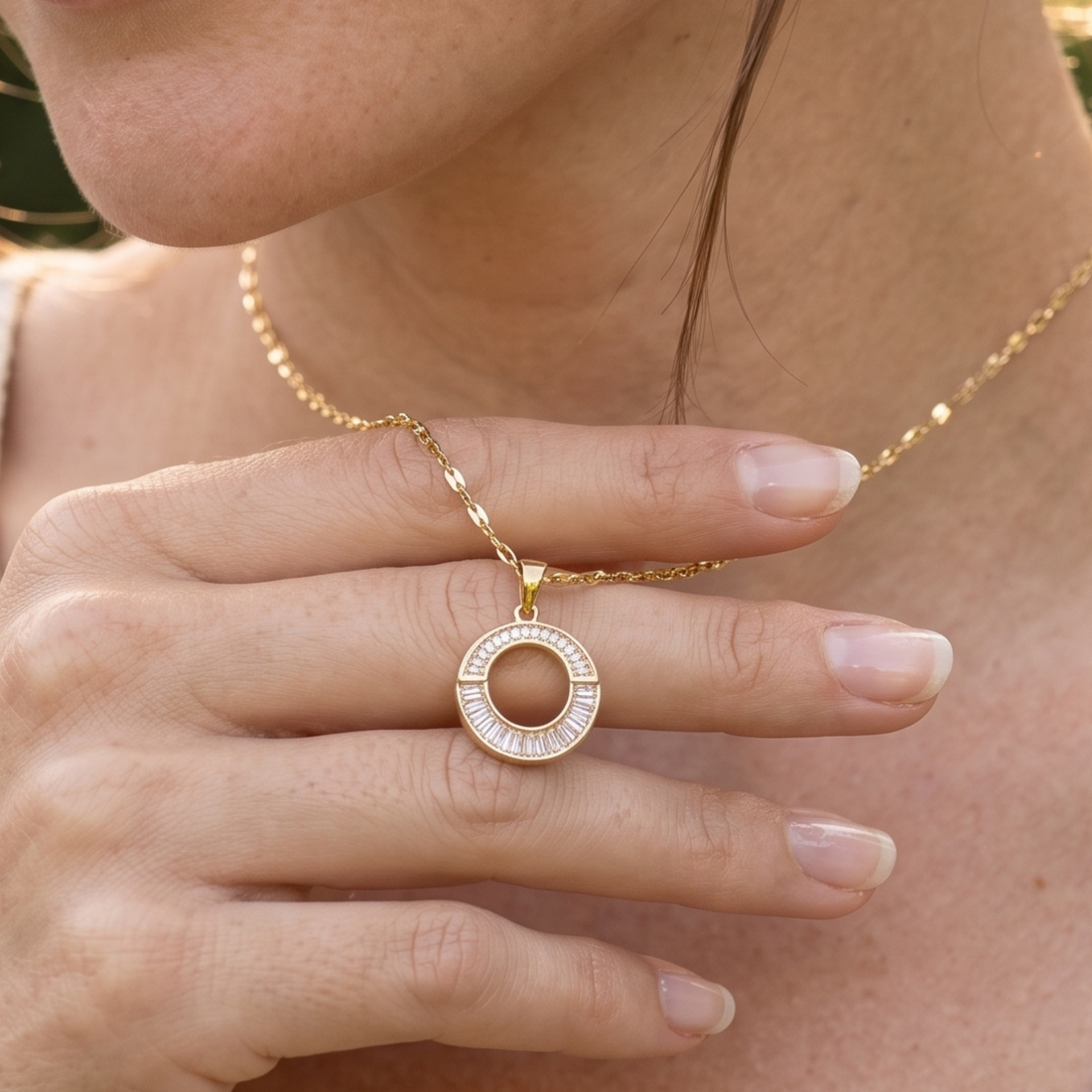 A close-up lifestyle shot of a model wearing a delicate gold chain necklace with a circular pendant. The pendant features a gold outer ring set with small clear stones, held gently by the model's hand.