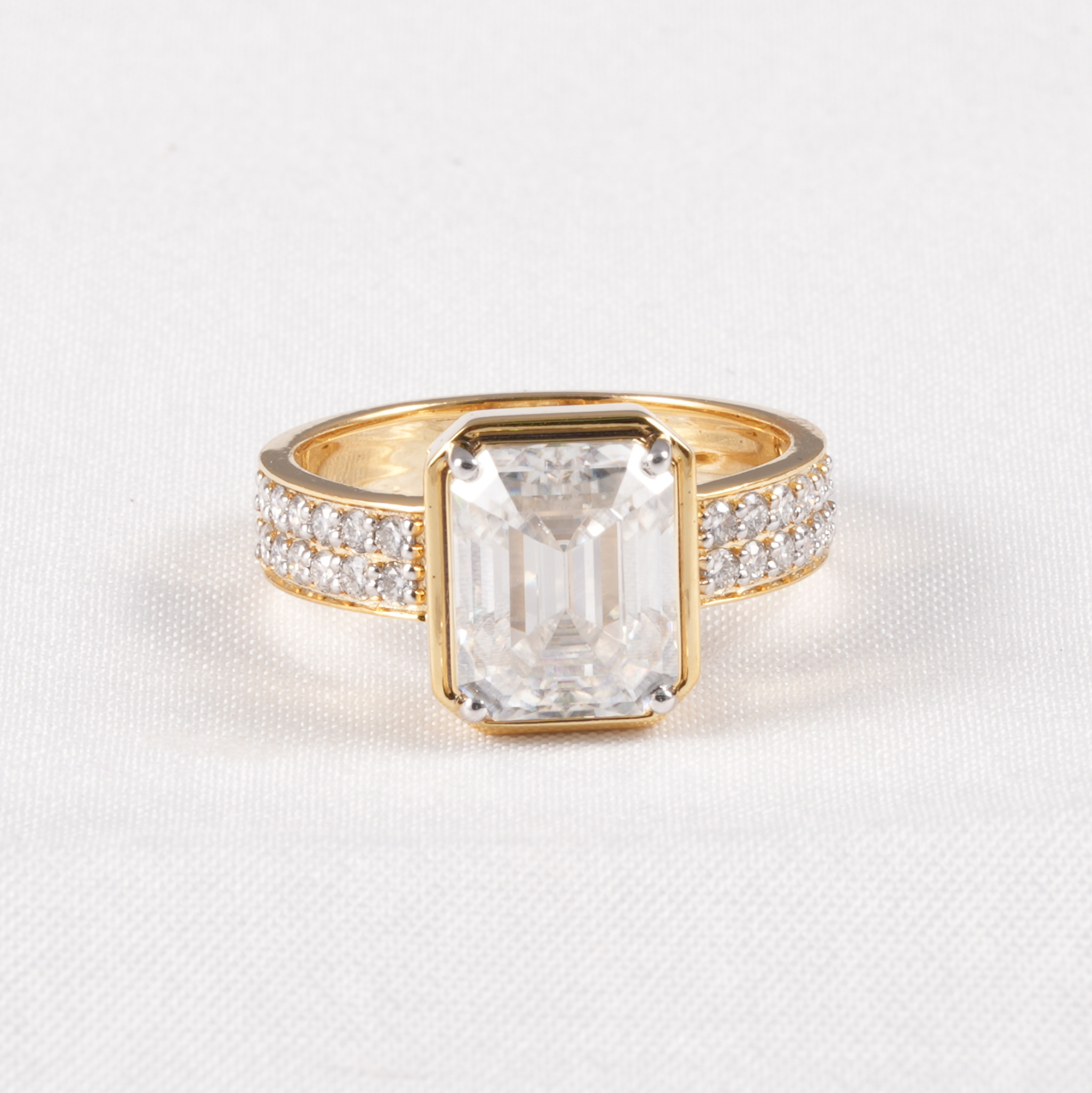 A yellow gold ring featuring a large rectangular emerald-cut clear center stone, flanked by pavé-set clear stones on the band, shown on a textured white background.