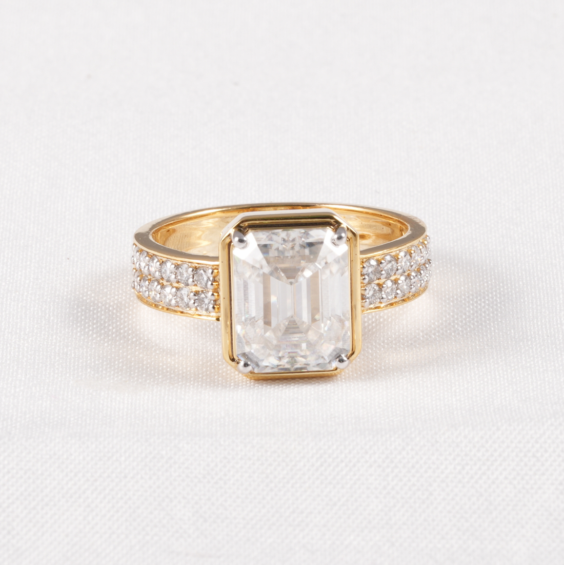 A yellow gold ring featuring a large rectangular emerald-cut clear center stone, flanked by pavé-set clear stones on the band, shown on a textured white background.