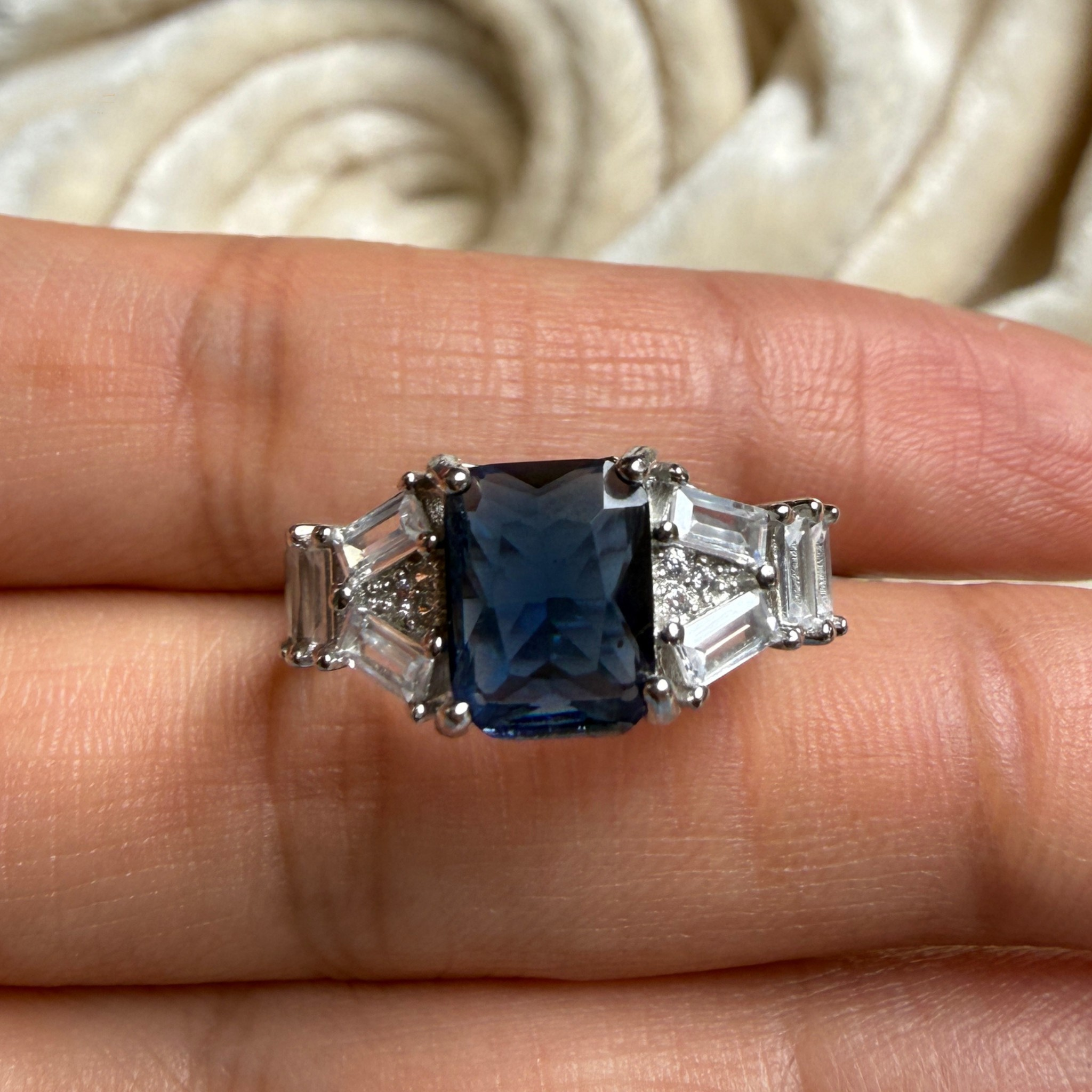 A silver-tone ring featuring a rectangular blue sapphire center stone with baguette and round side stones, shown worn on a finger.