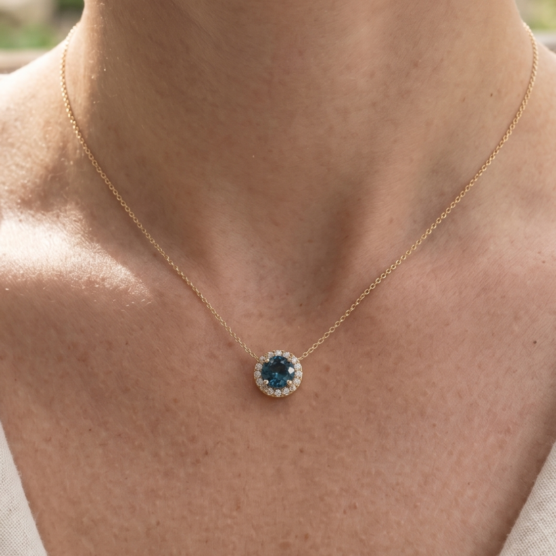 A close-up of a person's neck and upper chest wearing a delicate gold chain necklace with a round pendant featuring a deep blue center stone surrounded by small clear stones.