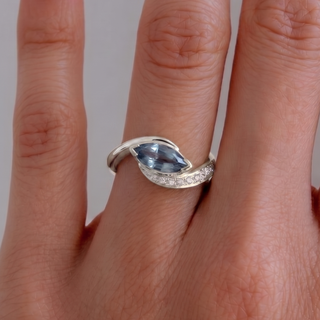 A silver ring with a marquise-cut light blue gemstone and a row of small clear stones, shown worn on a finger.