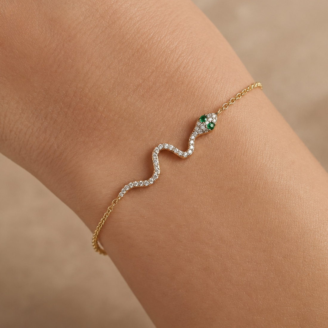 A delicate gold chain bracelet worn on a wrist, featuring a wavy silver pavé section with clear stones and a green and gold snake head at the end.