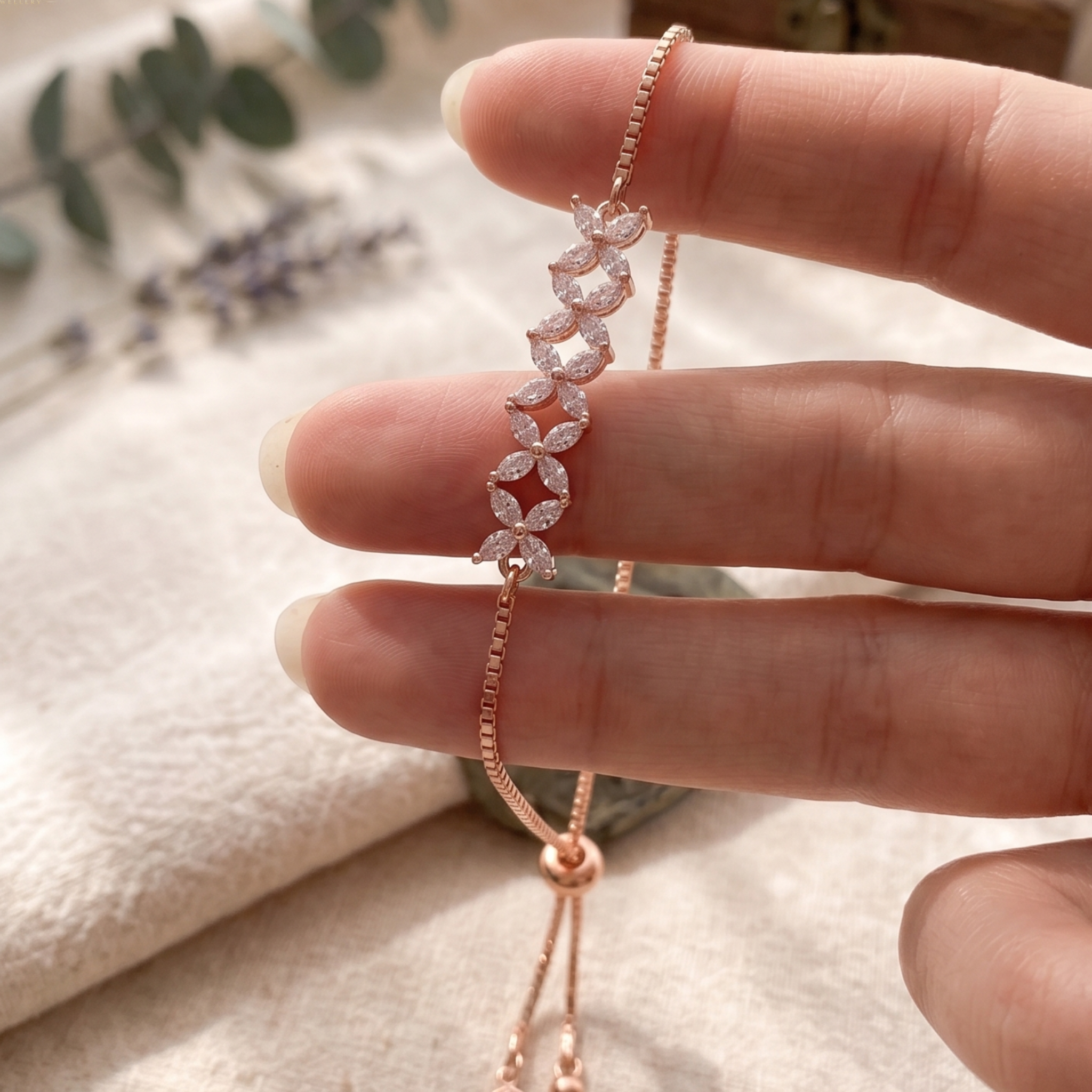 A close-up of a hand holding a delicate rose gold chain bracelet with a floral leaf motif paved with small clear stones, set against a soft beige fabric background with blurred greenery.