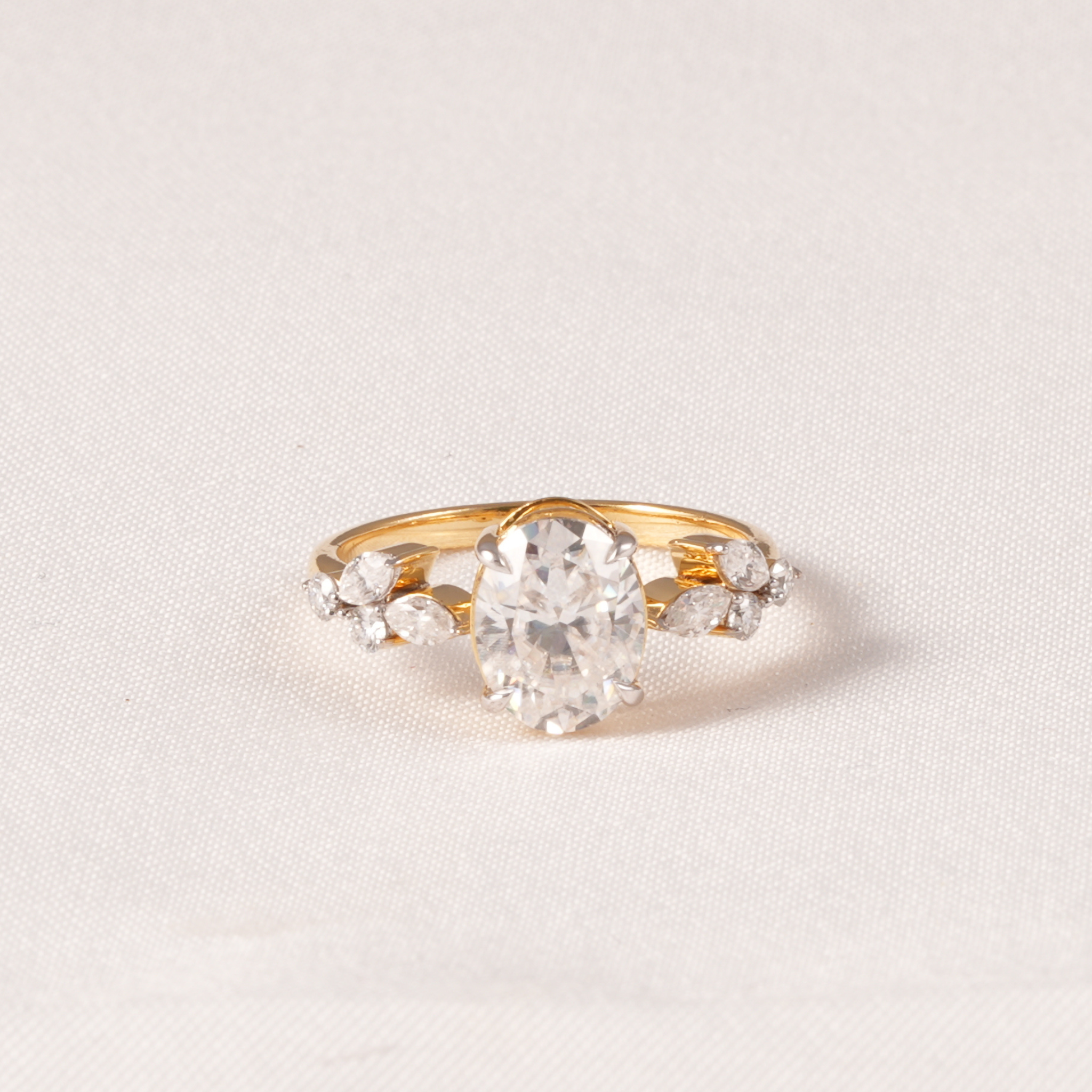 A yellow gold ring featuring a large oval-cut clear center stone and smaller marquise-cut side stones, photographed on a light beige textured fabric background.