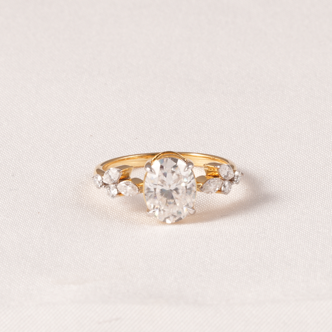 A yellow gold ring featuring a large oval-cut clear center stone and smaller marquise-cut side stones, photographed on a light beige textured fabric background.